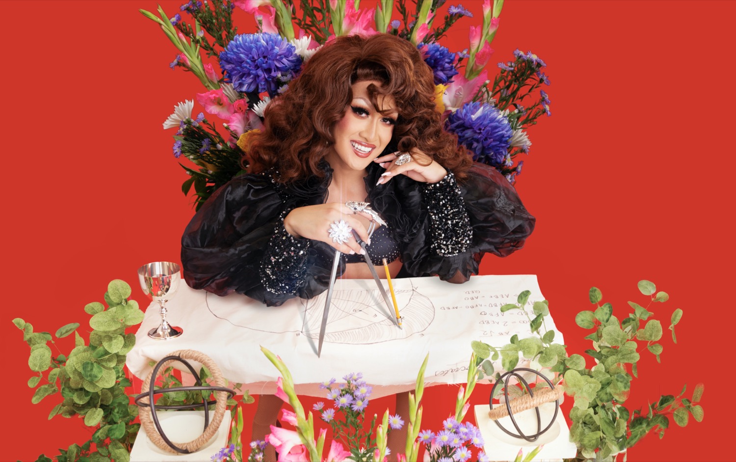 a white drag queen sitting at an extremely decorative desk with lots of bouquets of flowers around her. she's smiling at the camera sitting behind the desk holding a protractor over a sheet of paper, which has markings on it