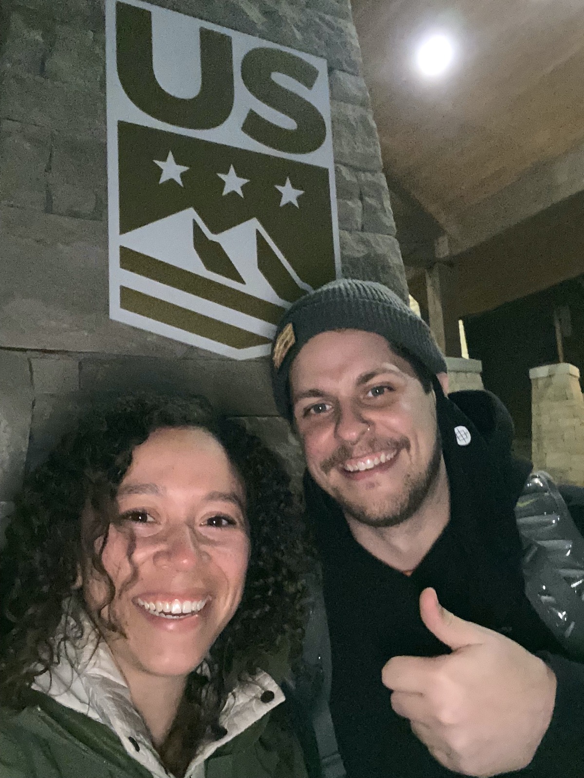 a white woman and white man posing for a selfie, smiling, the man giving a thumbs up. behind them on a wall is a u.s. ski team poster