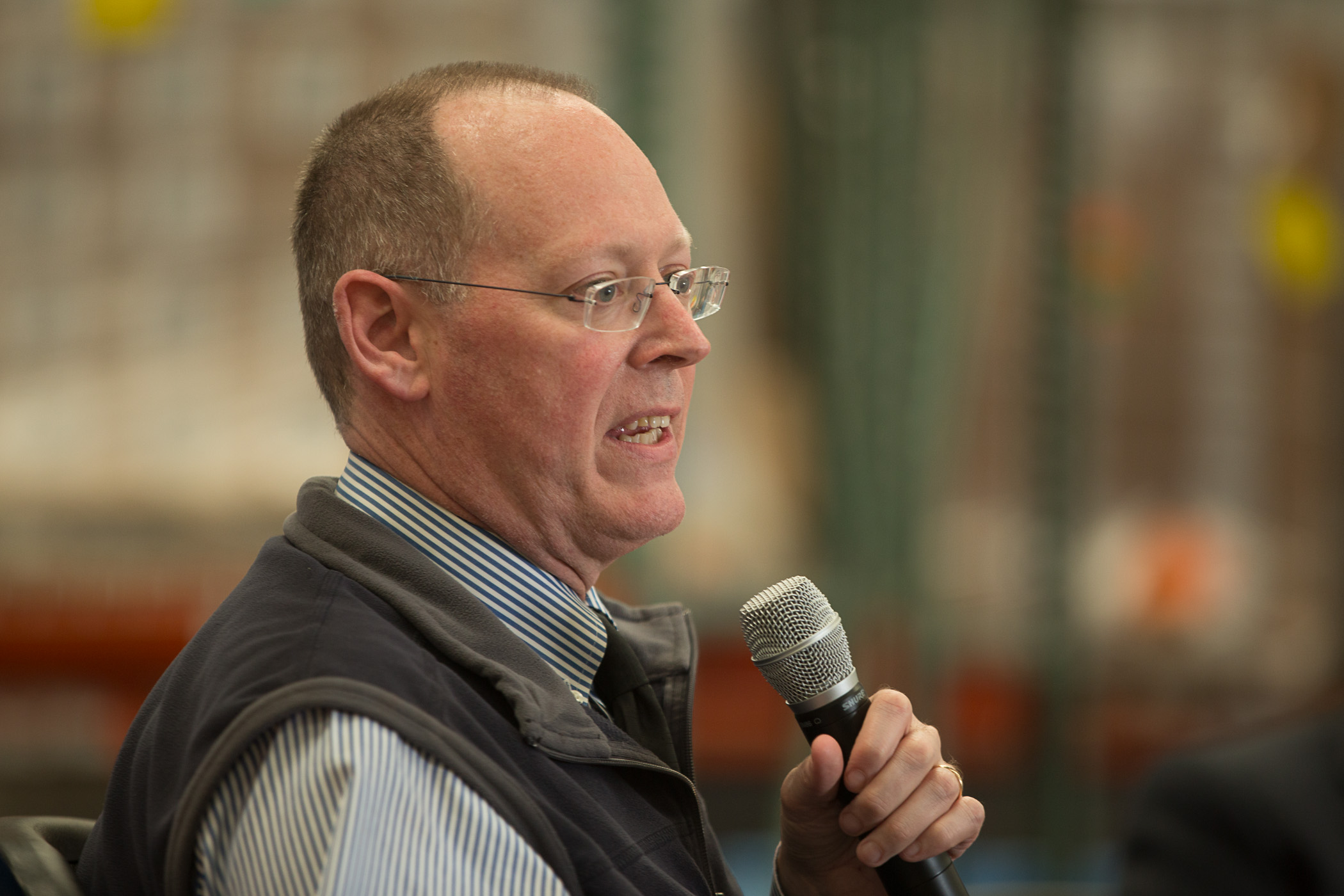 an older white man wearing glasses and a sweater vest speaking into a microphone