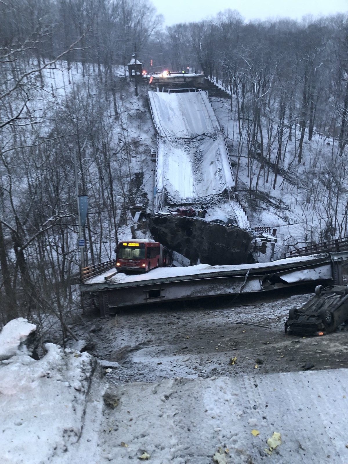 looking over the ledge of road of a collapsed bridge in snowy woods. bus is trapped under a large piece of debris