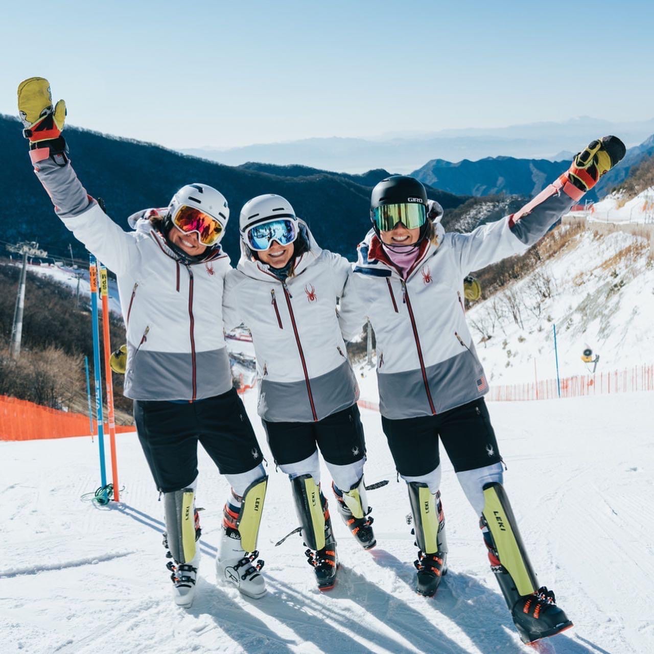three women in full ski gear with masks and goggles posing and smiling for the camera on top of a mountain