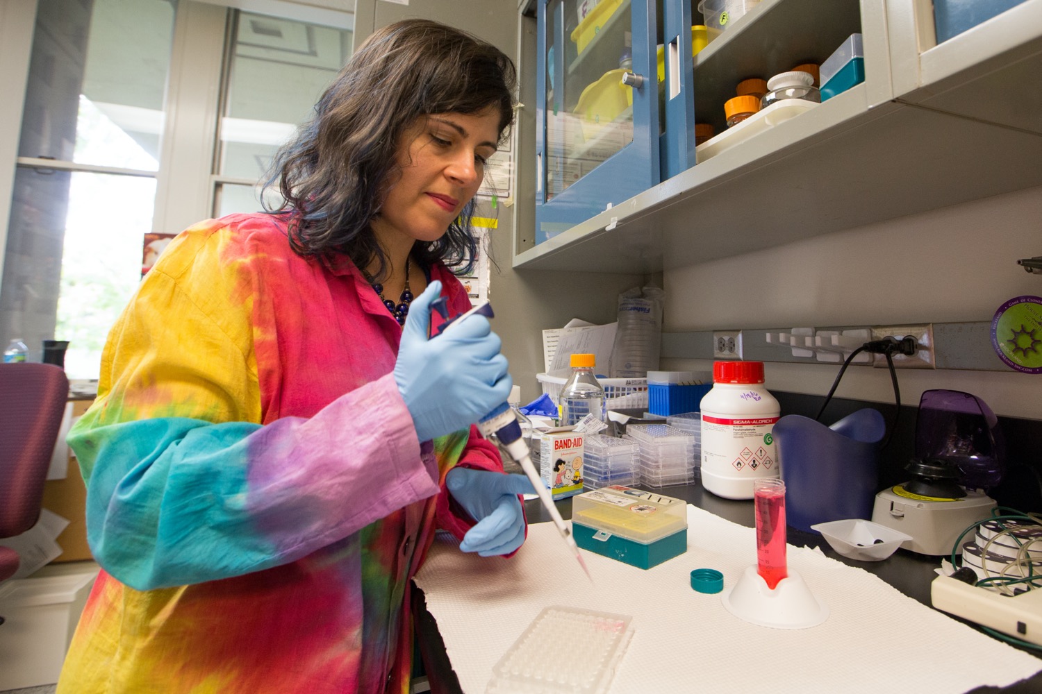 a woman in a lab wearing a multicolored lab coat holding a dropper over a container