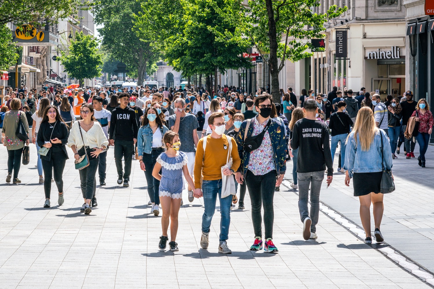 a large crowd on a bright sunny street. some people are wearing masks, most are not