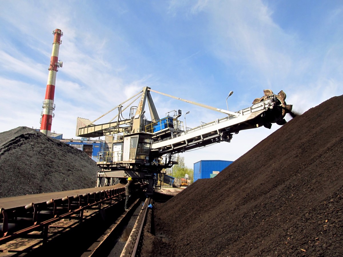 A large dark pile of goal rises in the foreground of the right side of the image, while a large machine with a boom like a crane rests in the background along a track. The machine, called a stacker, pours coal onto the pile on a sunny day with blue sky.