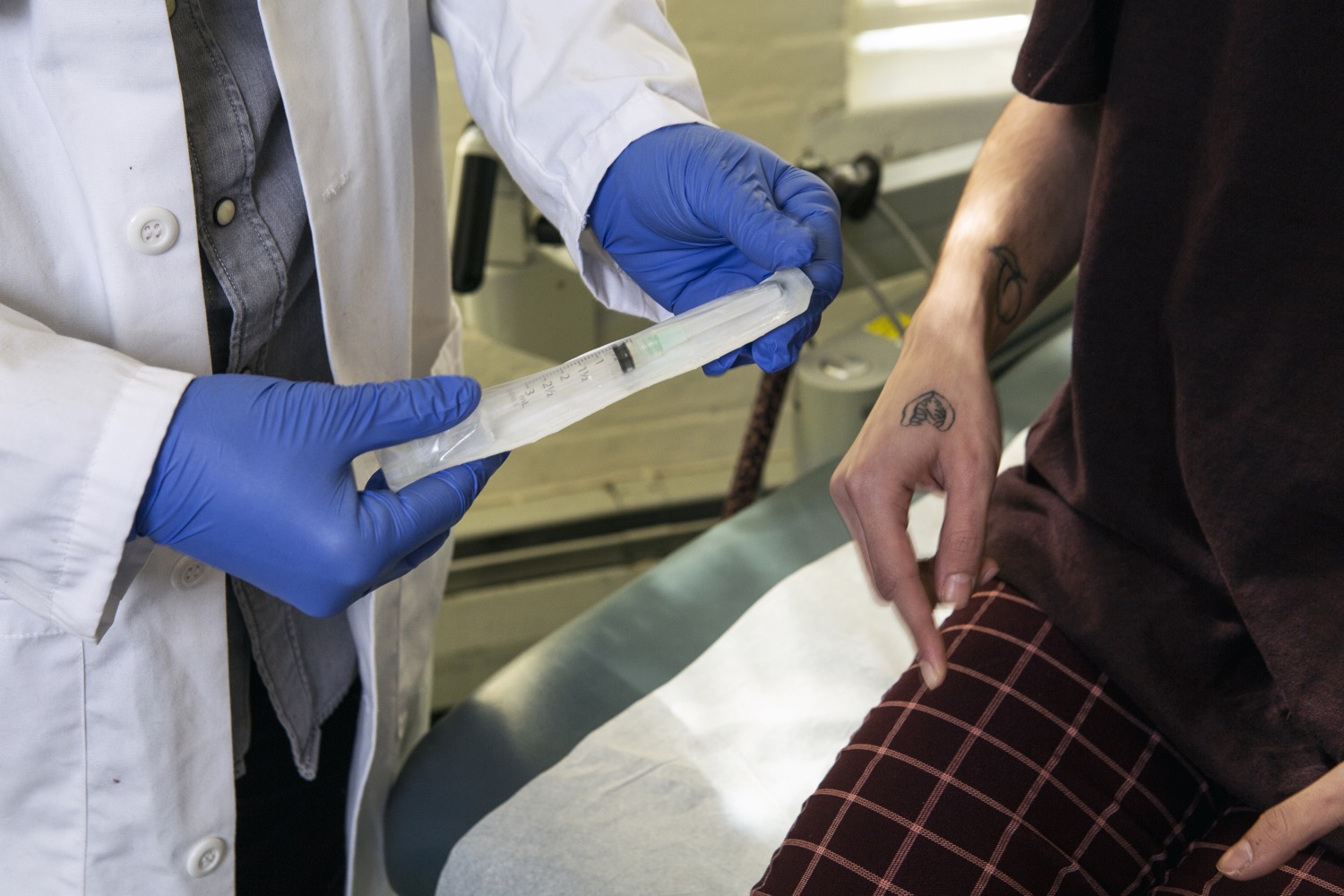 a doctor wearing blue gloves holding a syringe in a plastic bag next to a patient. all you can see are their torsos and legs