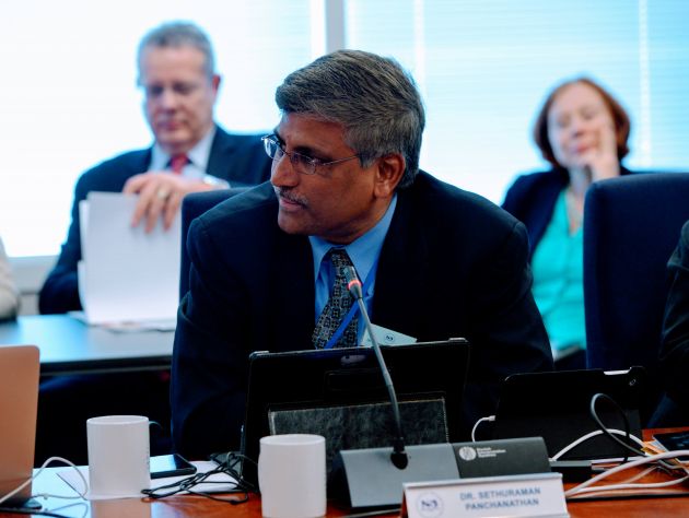 an older indian man in a suit tie in a meeting room sitting a desk, with an older white man and woman, also in suits, sitting behind him. a small microphone is in front of him as he looks to his right