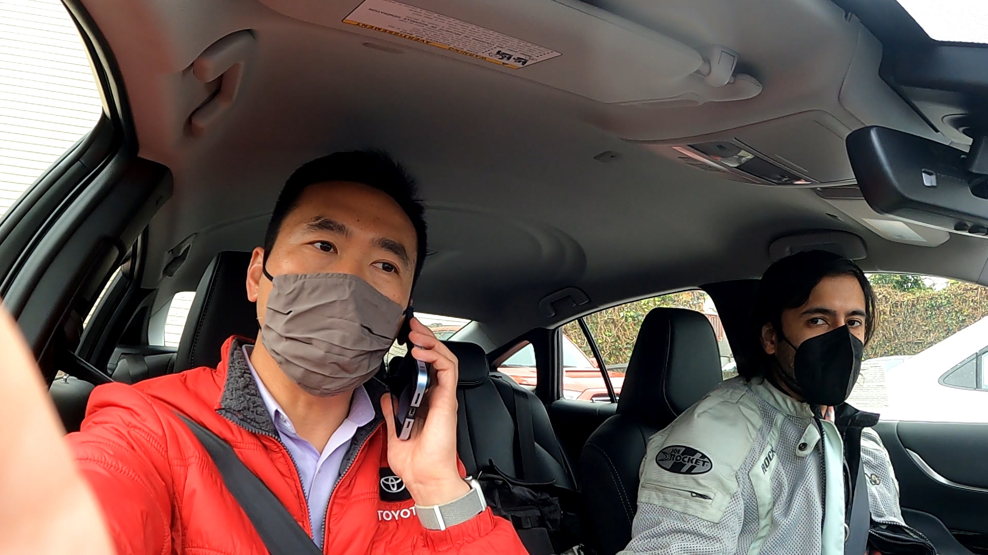 a selfie taken by a toyota representative in the passenger seat wearing a red toyota jacket and a man in the drivers seat, both wearing masks looking at the camera