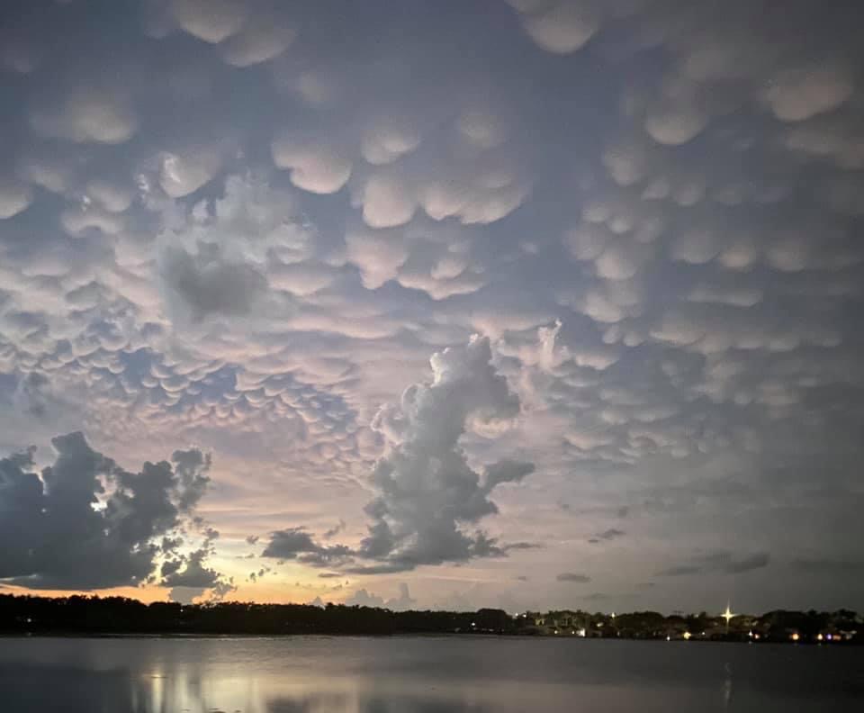 a landscape shot of a moody, dramatic sky, dotted by bulbous small clouds, jagged longer ones and a mild haze in the background. the color of the sky is darker, haunting, and sublime. the sun is setting, casting a reflection on the body of water in the foreground