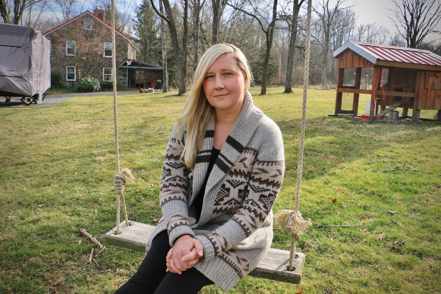 a white blonde-haired woman sitting on a swing outside looking pensive at the camera. behind her is grass, a shed, and a house