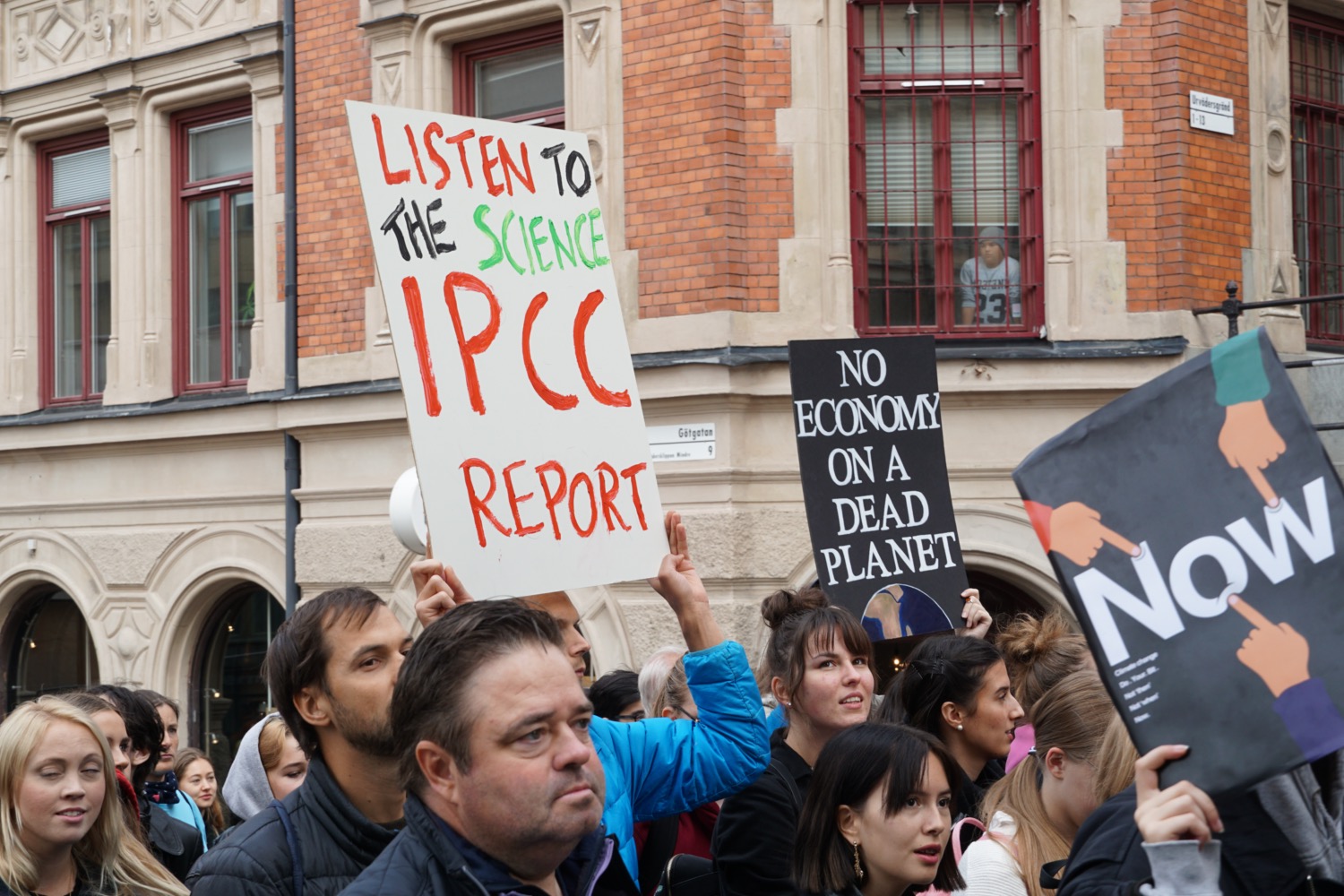 a crowd of protesters outside. three signs are being held up, they read "listen to the science ipcc report", "no economy on a dead planet", and "now""