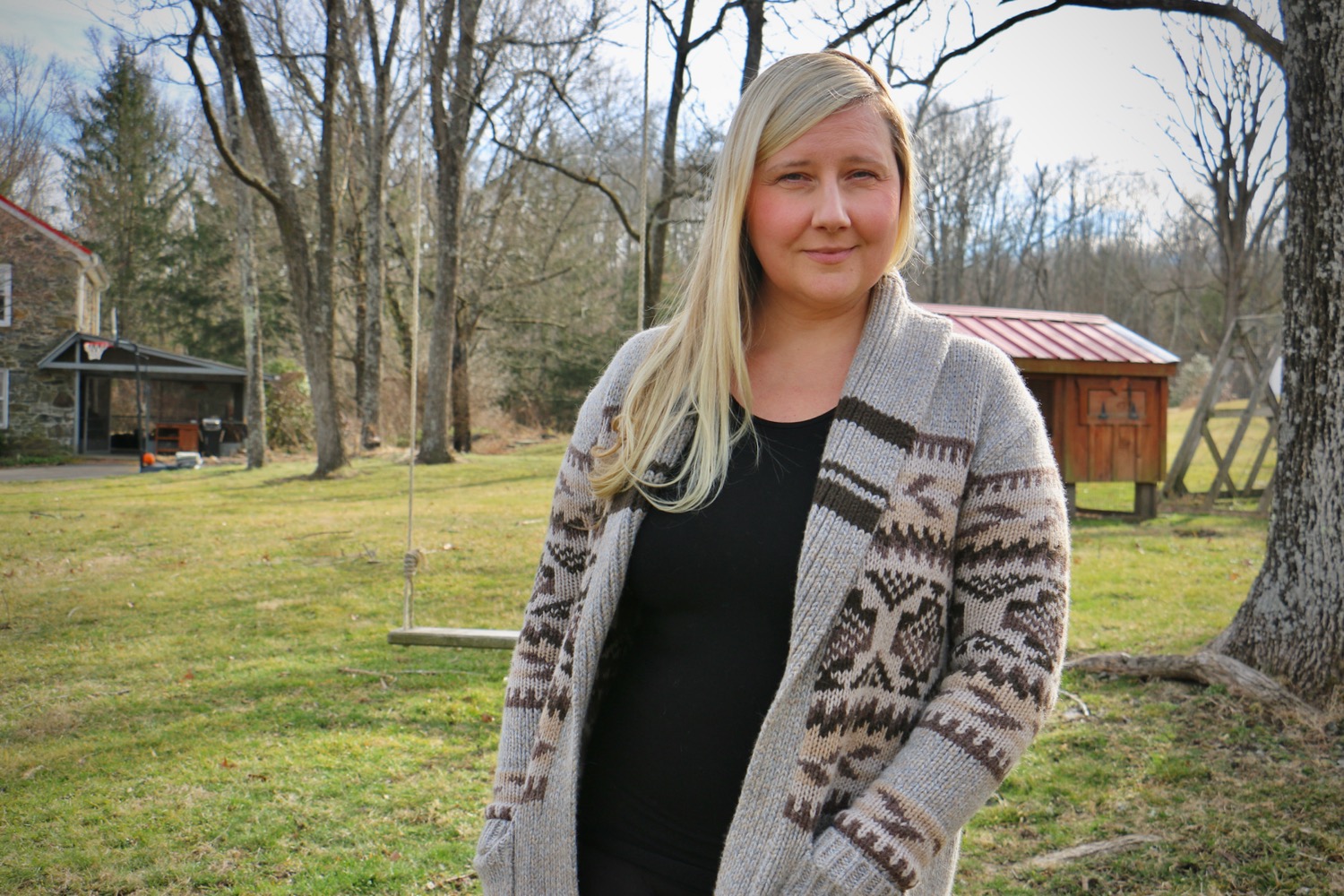 a white blonde-haired woman outside looking pensive at the camera. behind her is grass, a shed, and a house