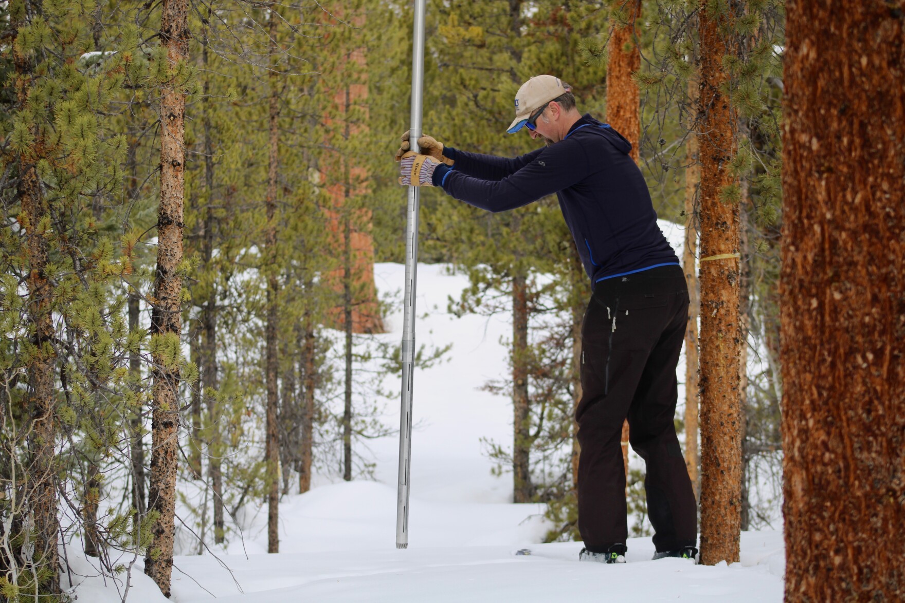 a white man in cold weather clothing and a baseball hat in a snowy forest. he holds a tall metal pole, an unknown amount of which is buried in the snow