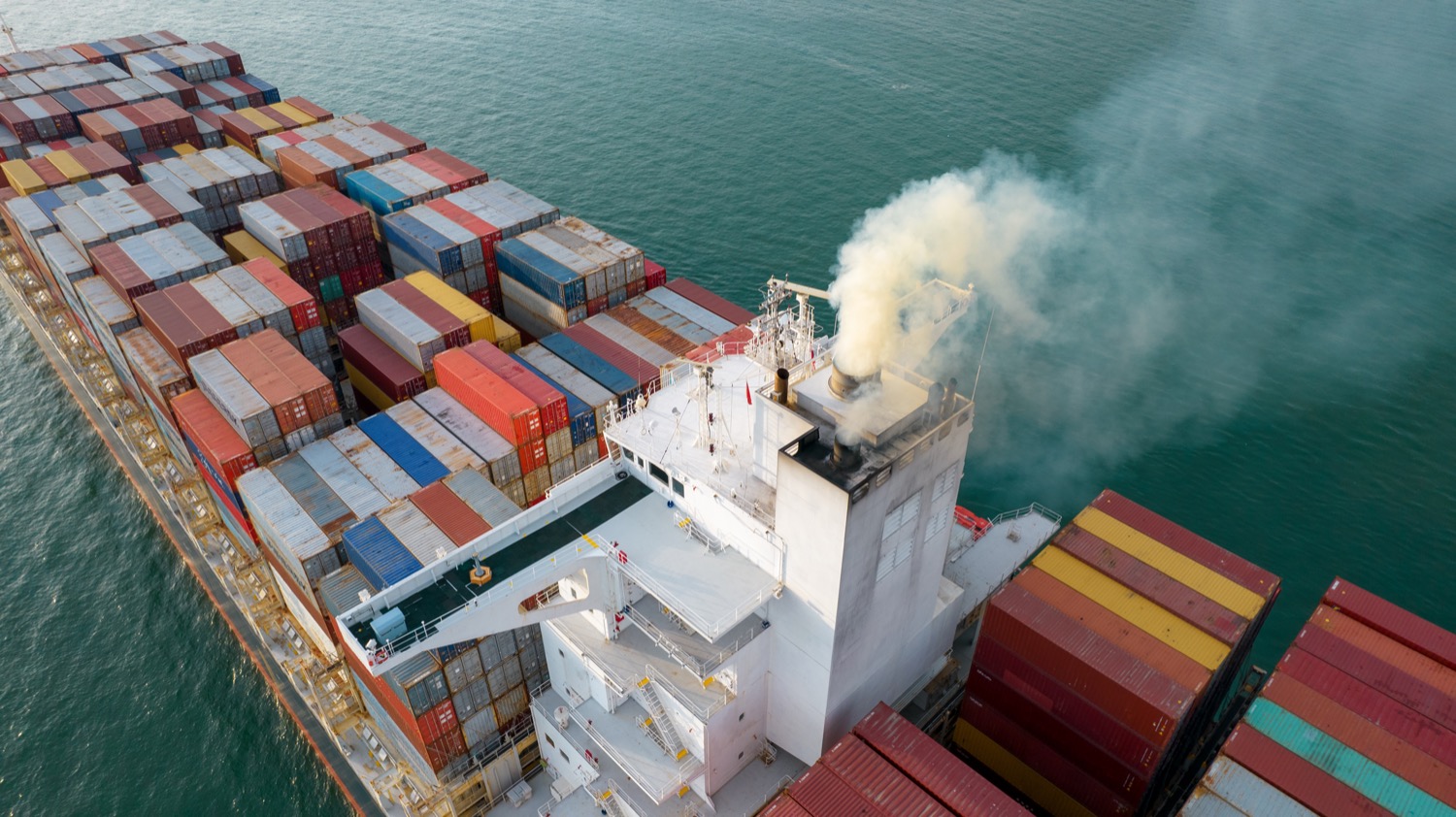 an aerial shot above a container ship loaded with containers on the ocean. its smoke stack emits smoke exhaust