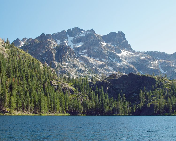 a landscape shot of a lake in the foreground, a lush pine forest behind it, and a dramatic, partially snow-covered jagged mountain in the background on a beautiful sunny day