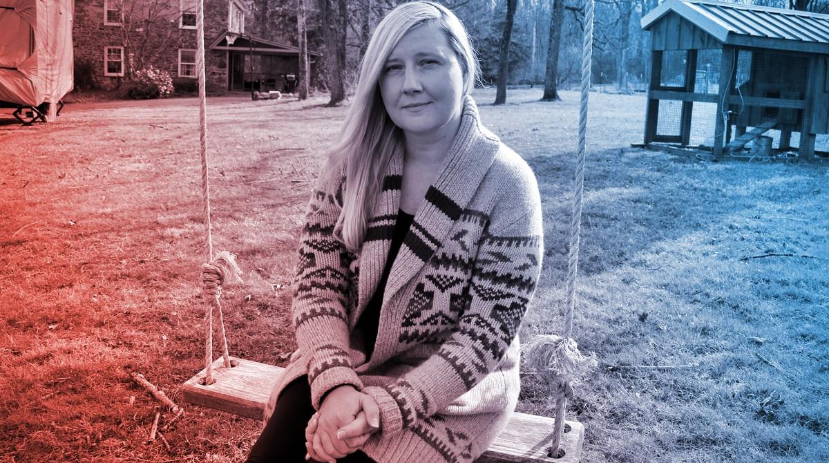 a white blonde-haired woman sitting on a swing outside looking pensive at the camera. behind her is grass, a shed, and a house