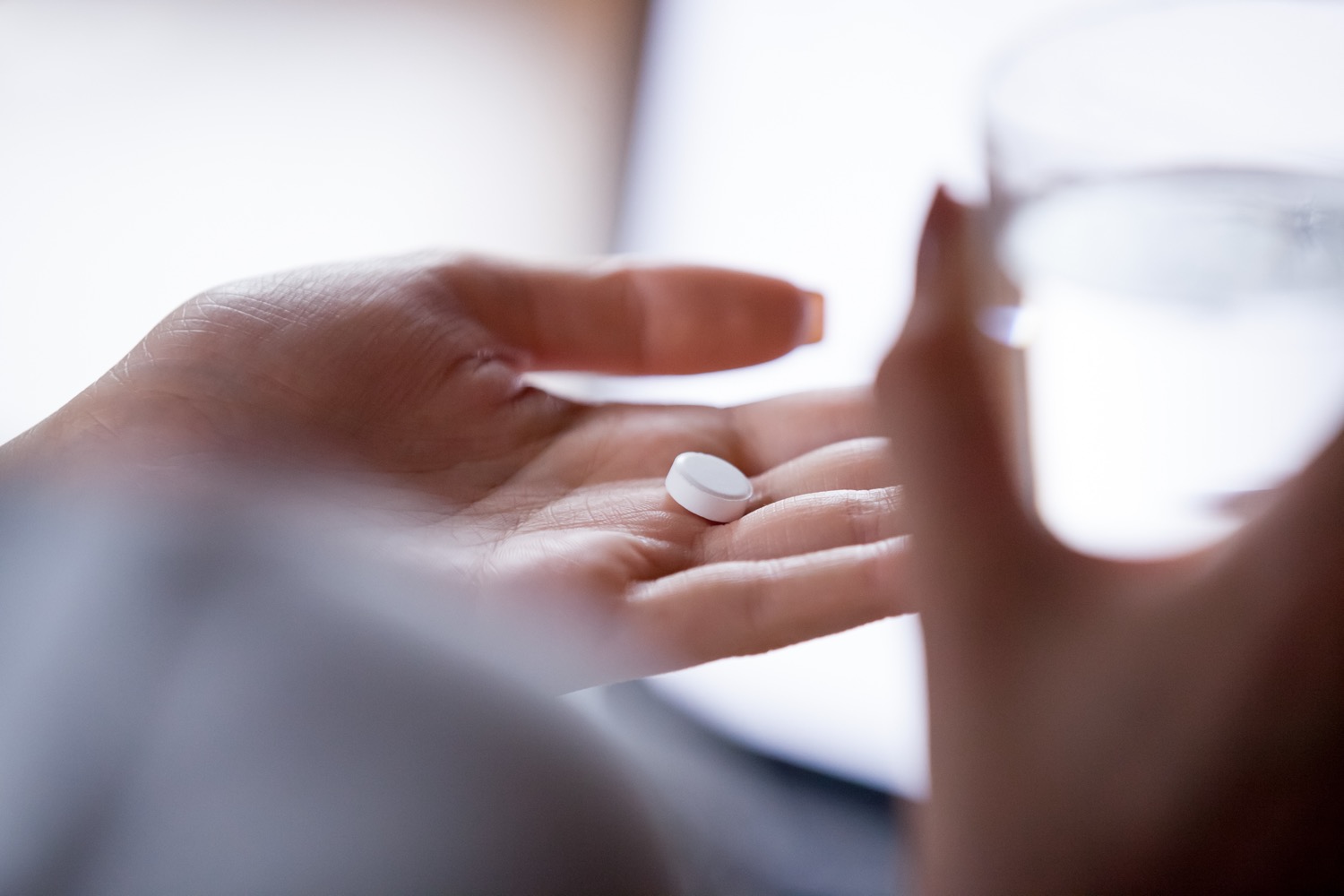 a close up of a white hand holding a small circular pill and a glass of water in the other hand