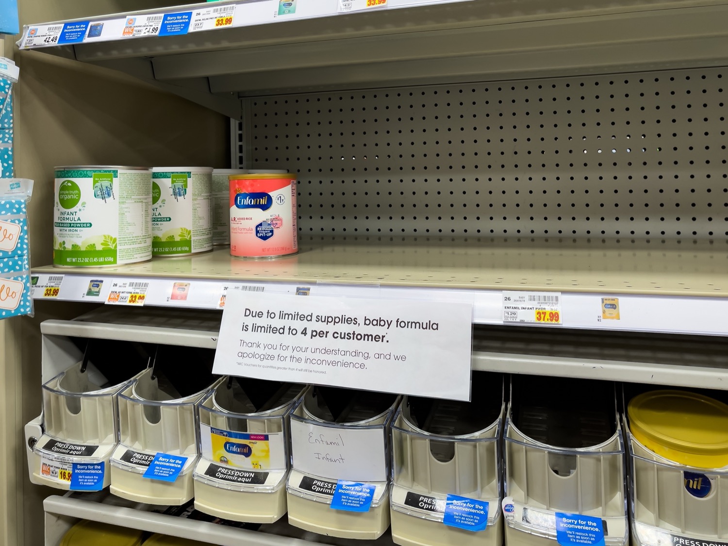 a nearly empty store shelf, on the left are only a few cans of baby formula. a small sign below the shelf reads 'due to limited supplies, baby formula is limited to 4 a customer. thank you for understanding and we apologize for the inconvenience."
