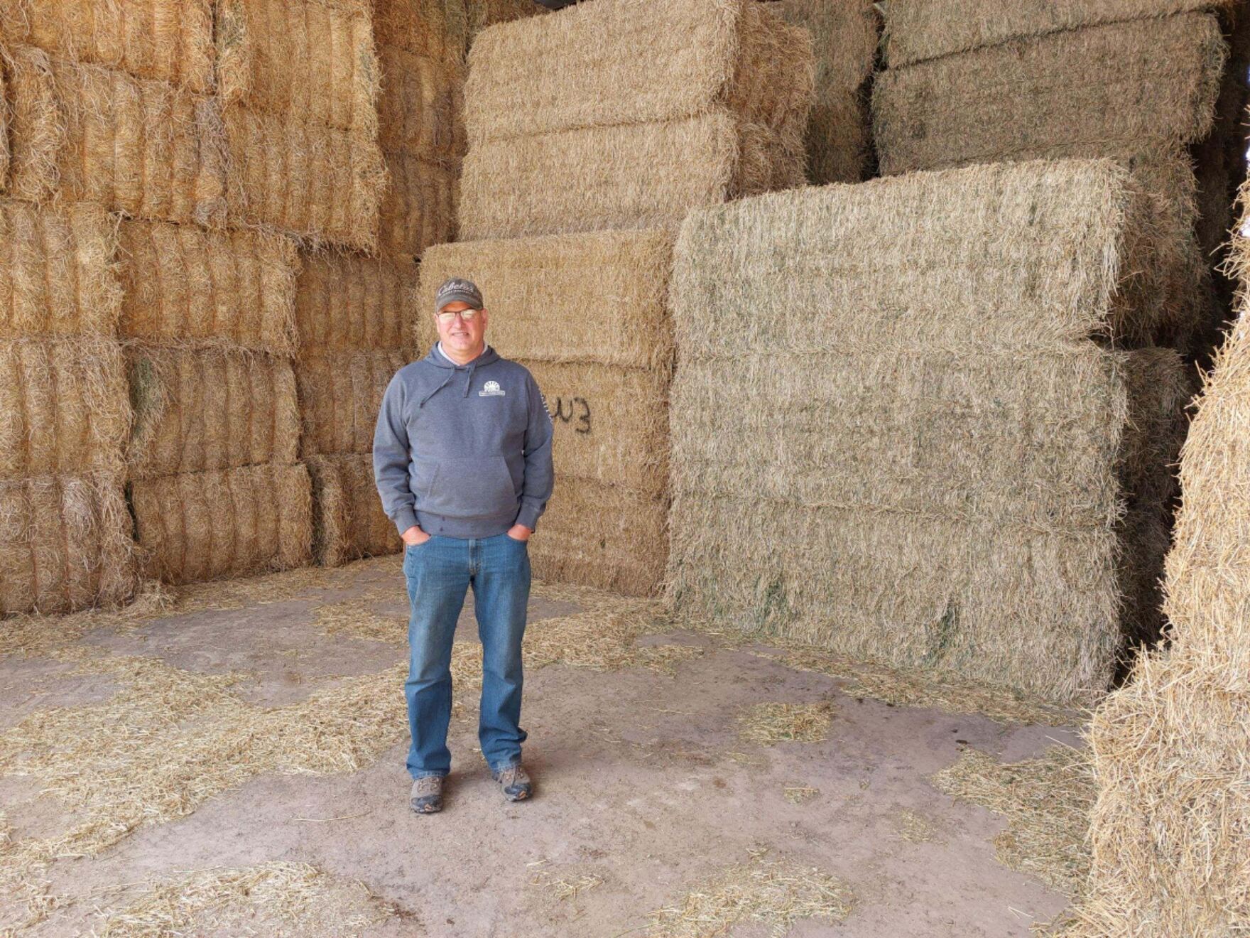 a man in a cap and sweater standing in front of bales of hay.