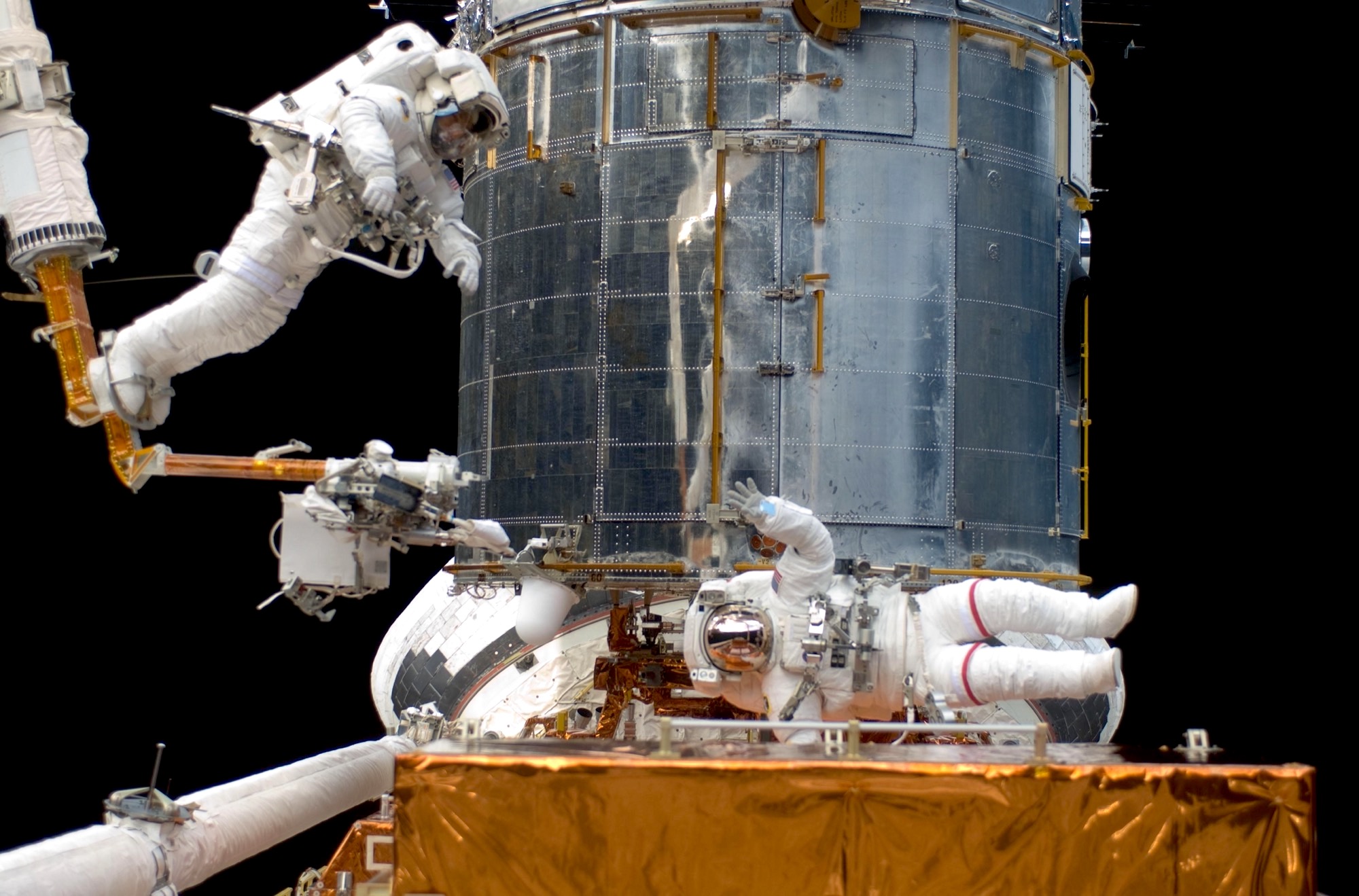two astronauts in space repairing the hubble space telescope, behind them. the telescope is far larger than they are. one of the astronauts wave at the camera