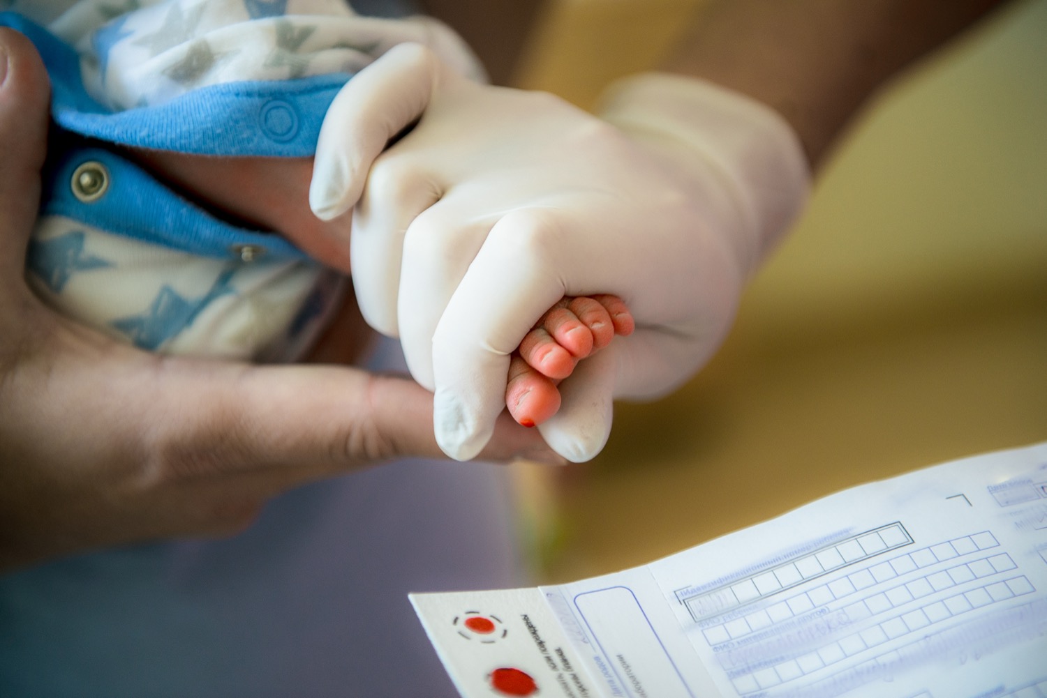 a medical gloved hand holding an infant's tiny foot, there's a small prick on the big toe, with some blood coming out so it can be caught on a blood test card held beneath it