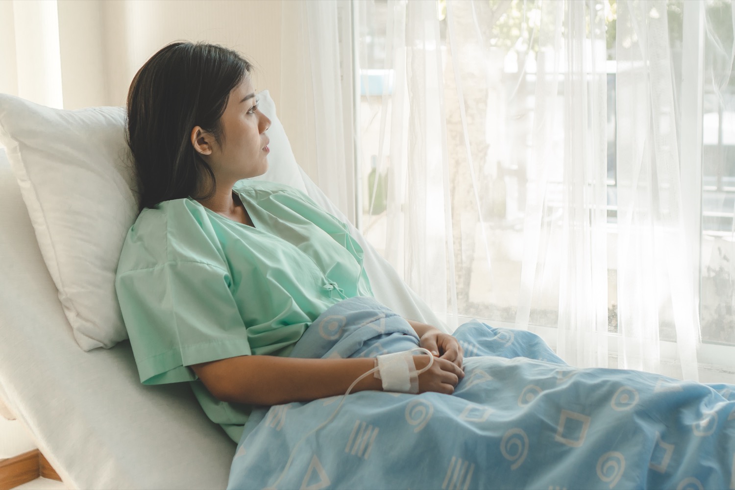 an asian woman on a hospital bed wearing a hospital gown sitting partially upright with her lower half covered by a blanket, looking out the window