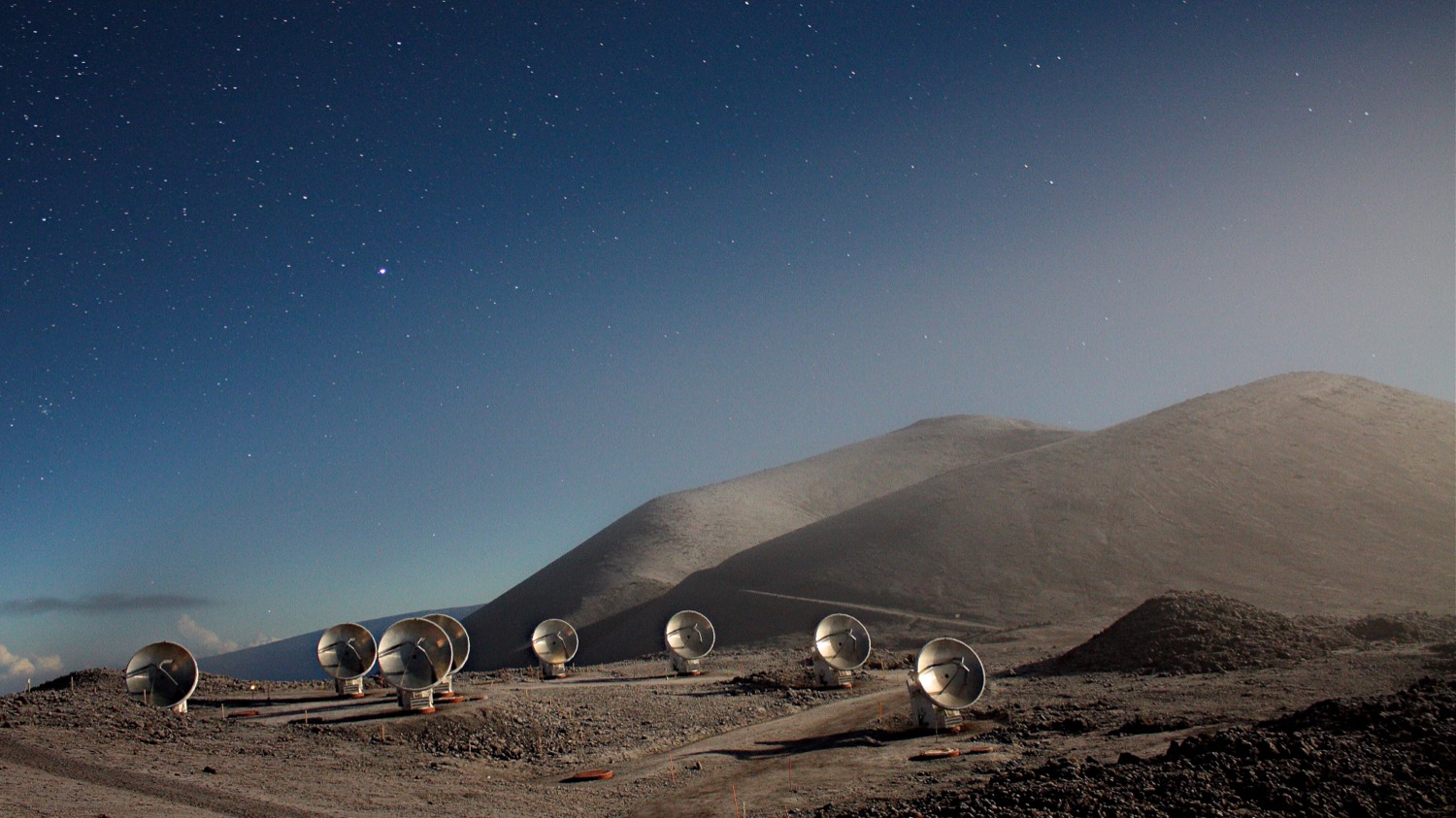 a group of telescopes with radio dishes among dunes and sitting under a dusk sky