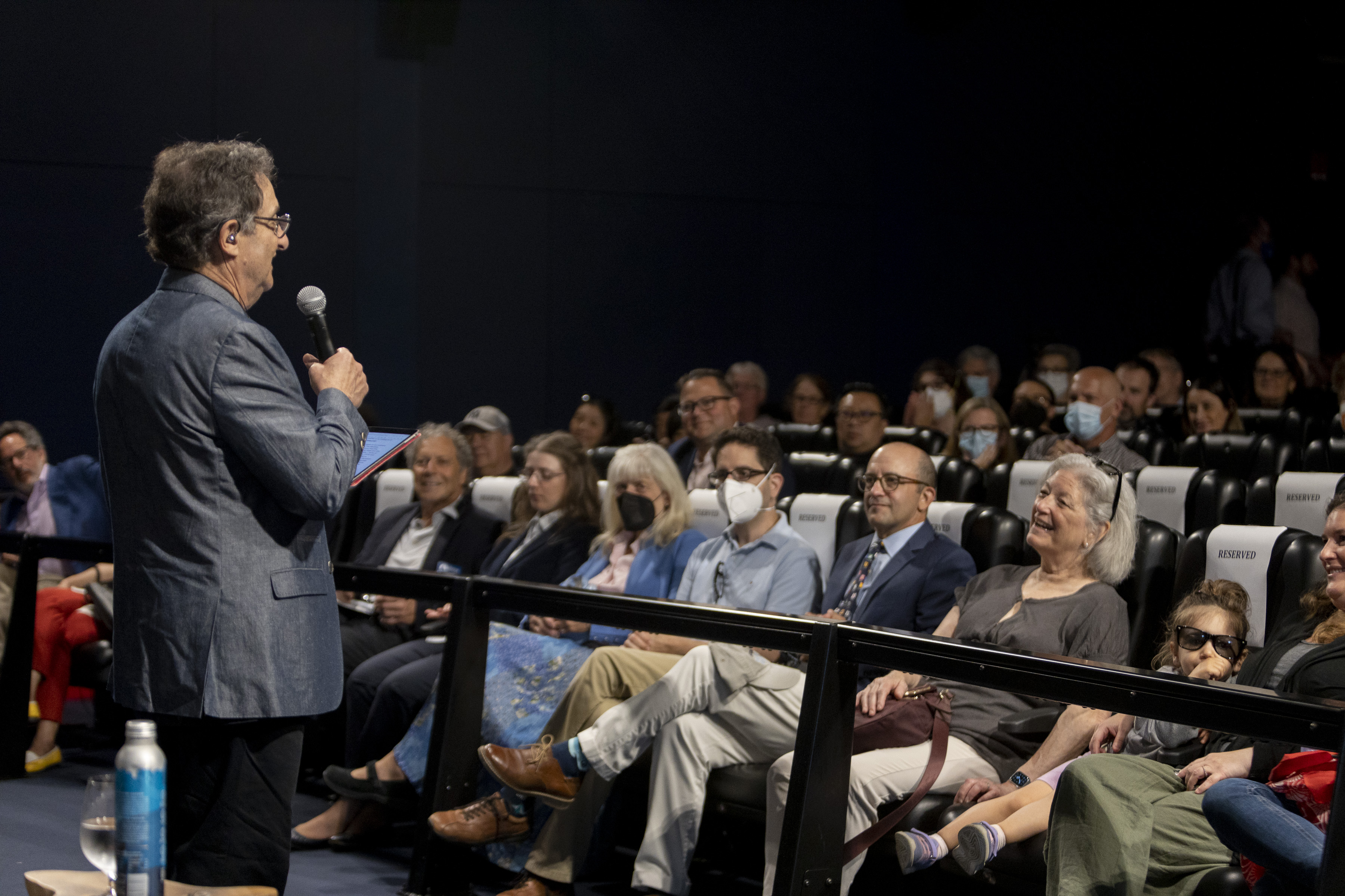 a picture taken behind ira flatow, wearing a suit jacket, speaking into a microphone addressing a smiling audience