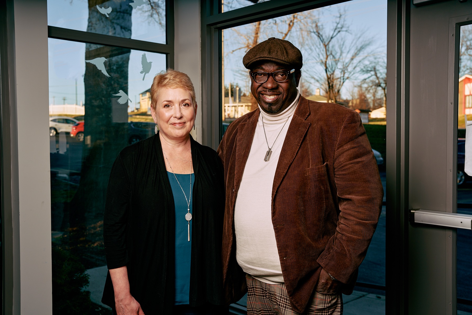 an older white woman with short blonde hair and an older black man wearing a newsy cap stand next to each other outside a building smiling at a camera