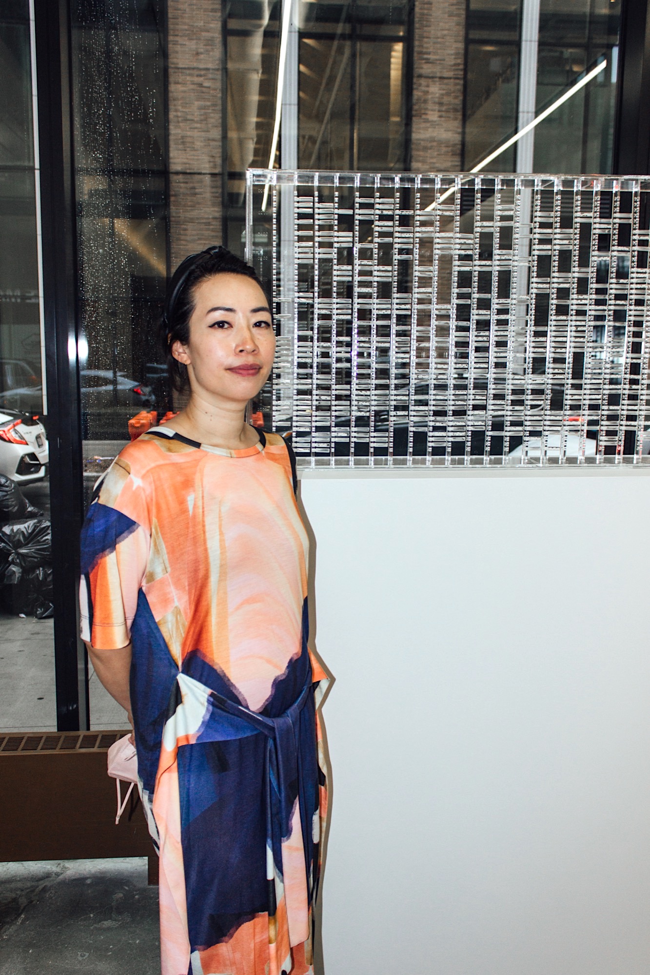 an asian woman wearing a flowy multicolored dress staring at the camera neutrally. she's standing beside one of her artworks, comprised of dozens of tiny see through tubes arranged in a display