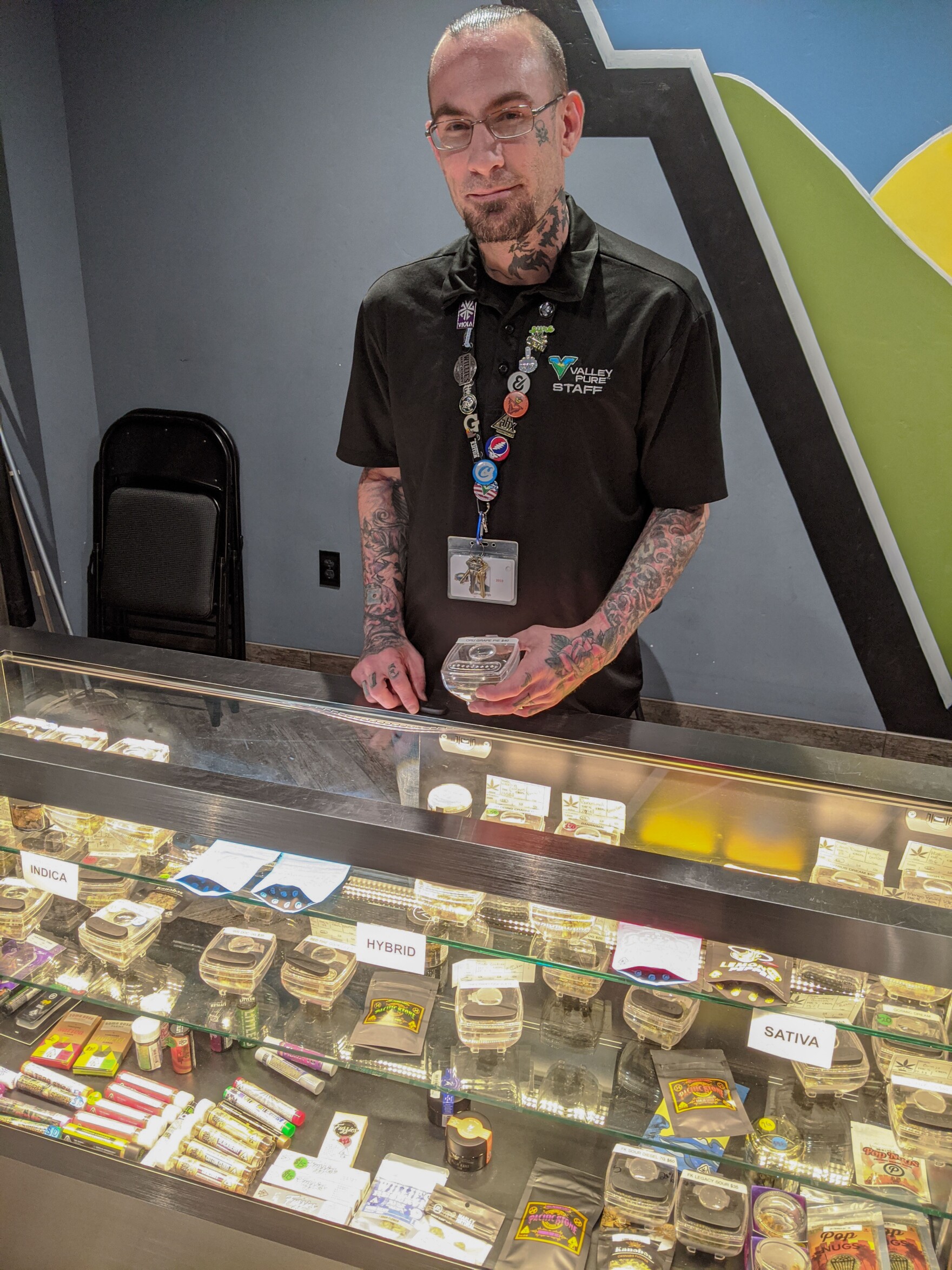 a smiling bald white man with tattoos stands behind a counter in a weed shop, the glass display case is filled with marijuana products
