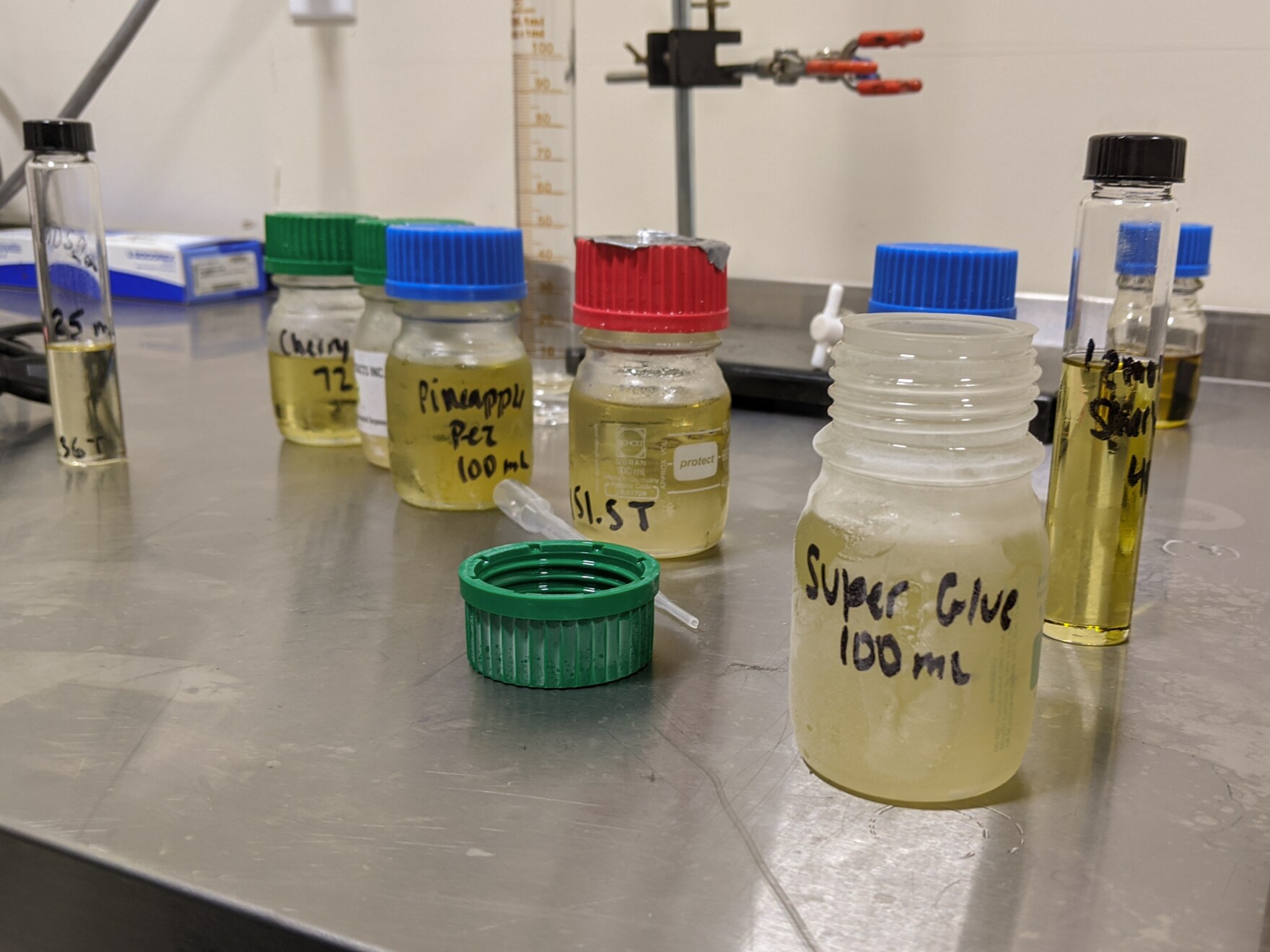 five lab bottles filled with a yellow liquid sit on a metal table in a lab