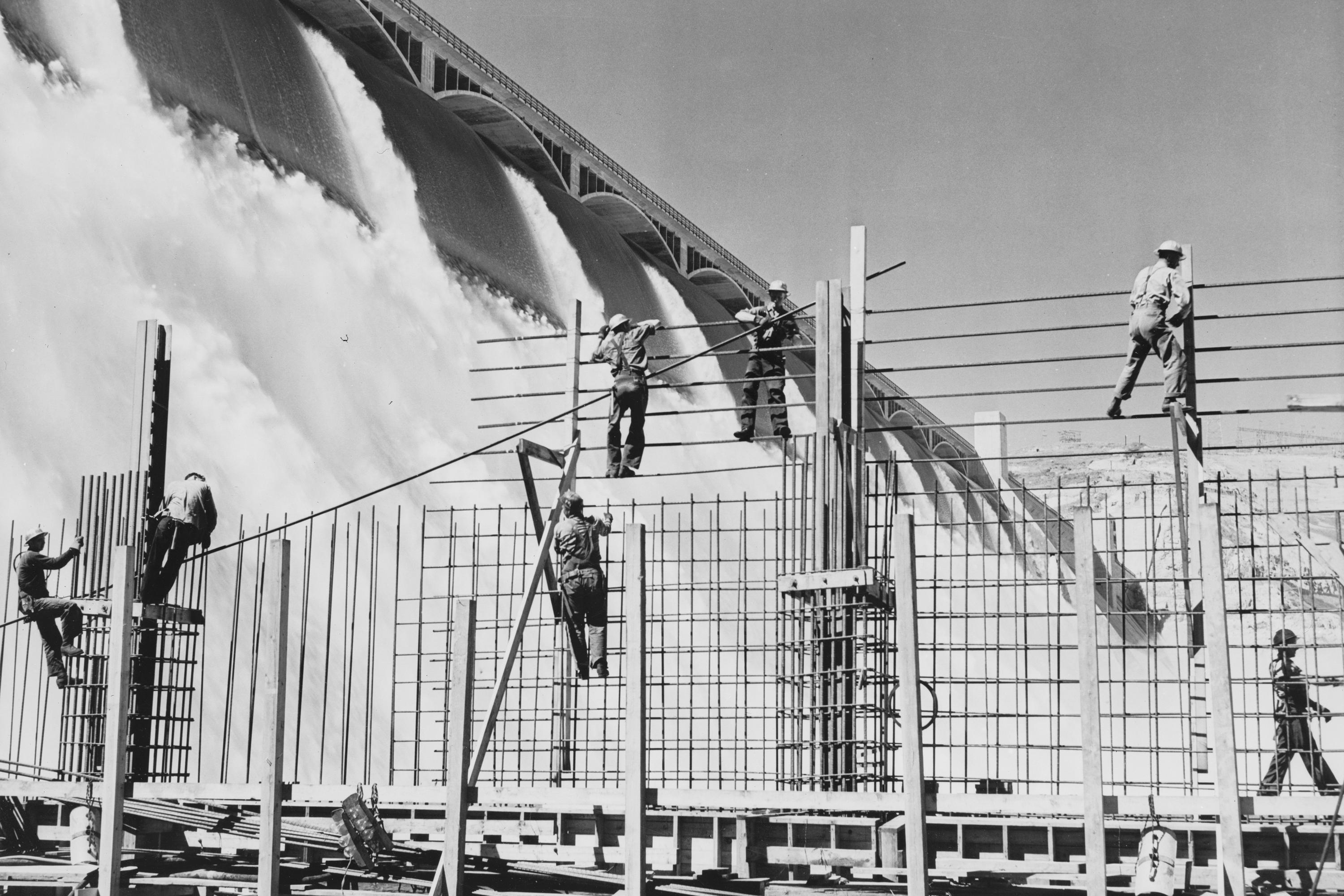seven men constructing scaffolding with a massive gushing dam behind them