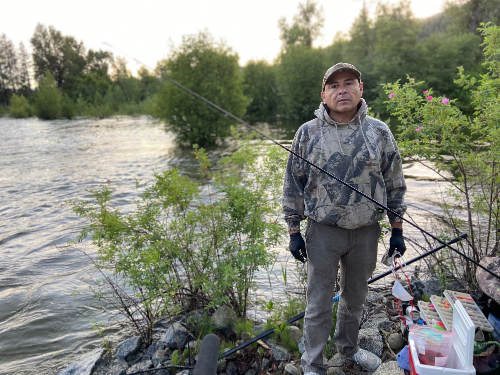 A man in a camo hoodie stands on a riverbank, surrounded by fishing gear.