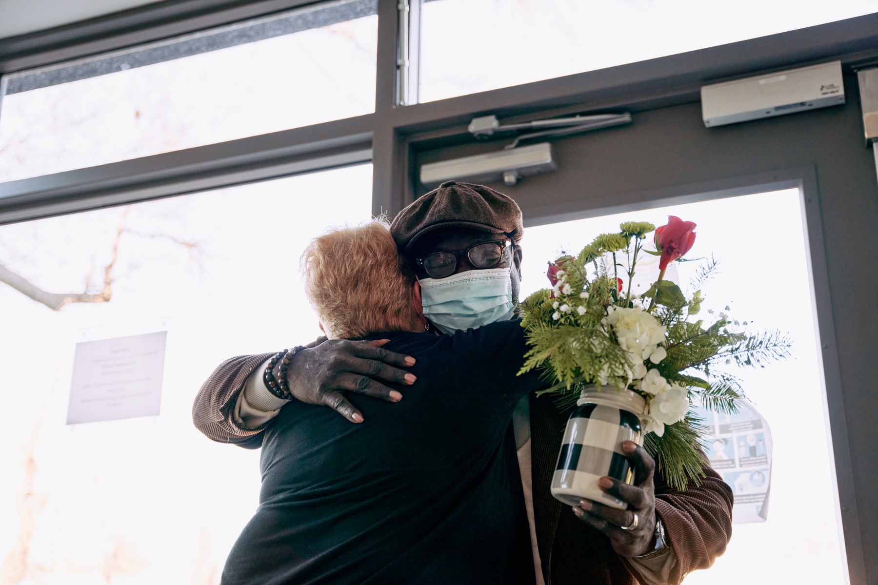 an older black man and an older white woman hug each other. the woman's back is to the camera and the man, wearing a mask, a newsy cap and glasses, is facing the camera, embracing the woman with one arm and holding a bouquet of flowers in his other hand