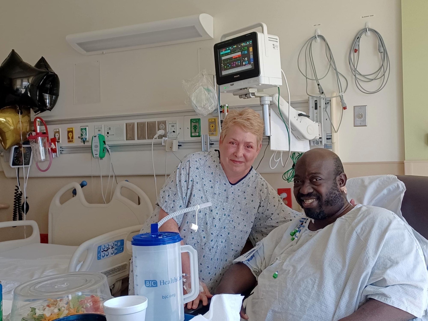 an older black man and an older white woman in hospital gowns in a hospital room smile at the camera. the man is sitting down while the woman is standing