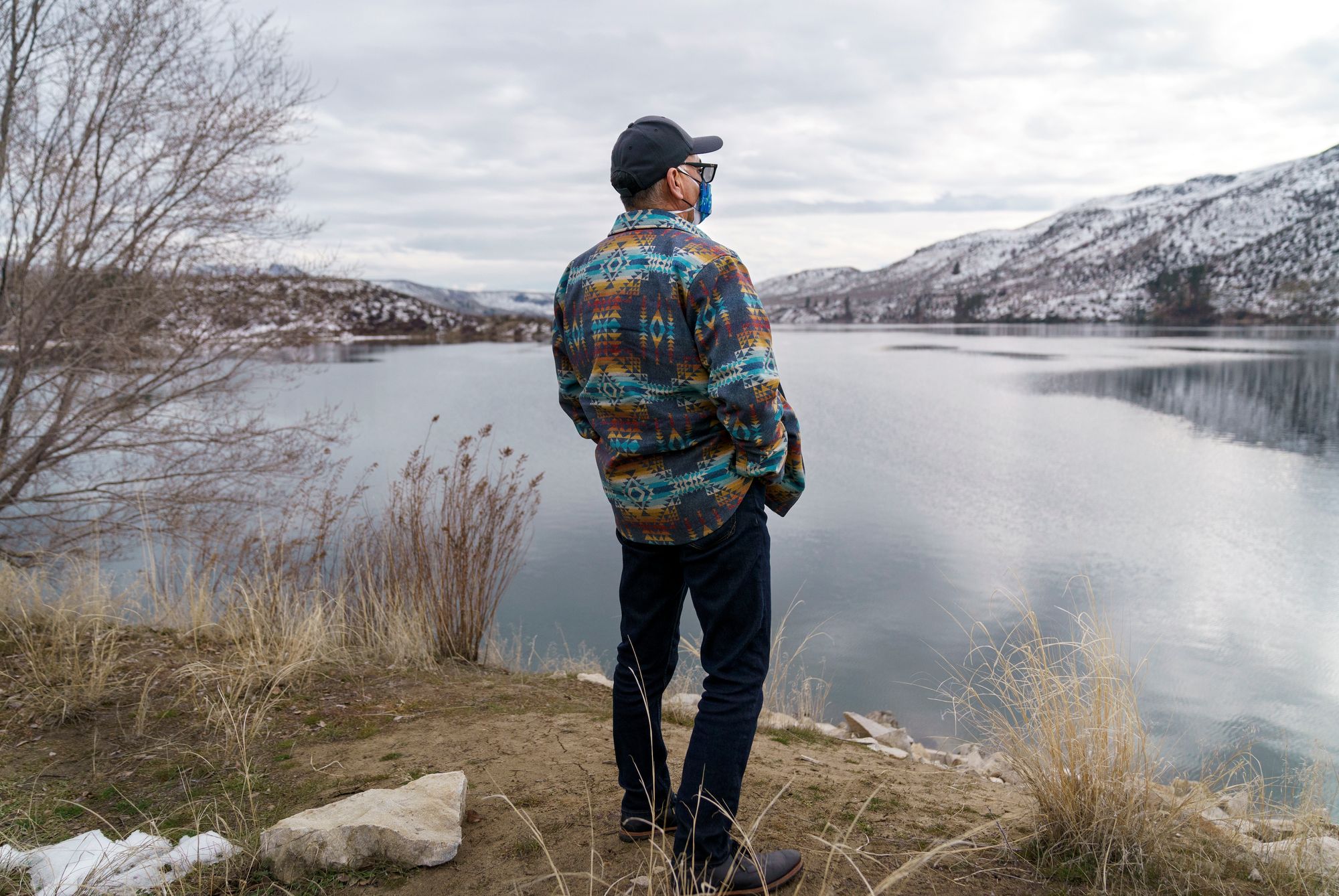 A man with his back to the camera looks over a calm body of water surrounded by snowy hills.