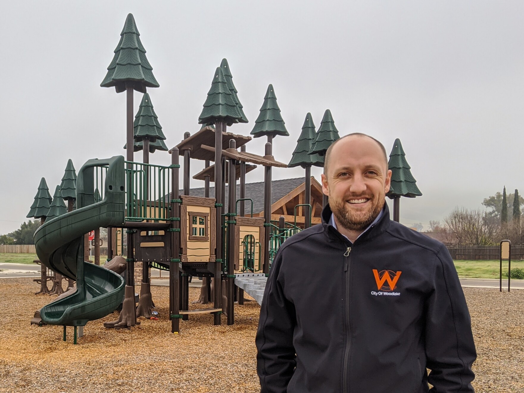 a smiling balding white man stands in front of a children's playground on an overcast day