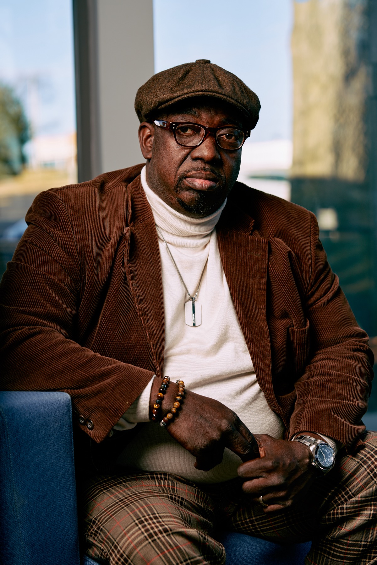 an older black man sitting on a chair wearing a newsy cap, looking pensively at the camera