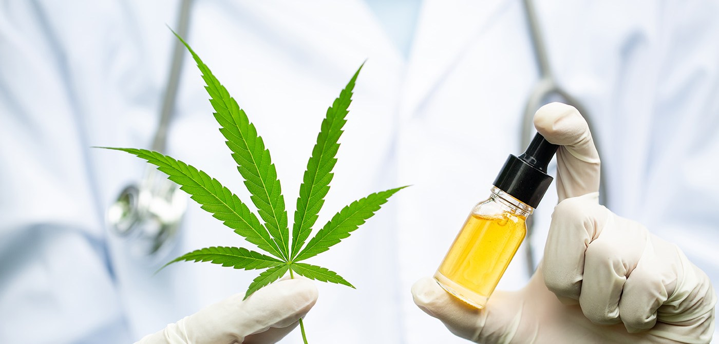 A doctor in a lab coat holding up a marijuana leaf and CBD oil.