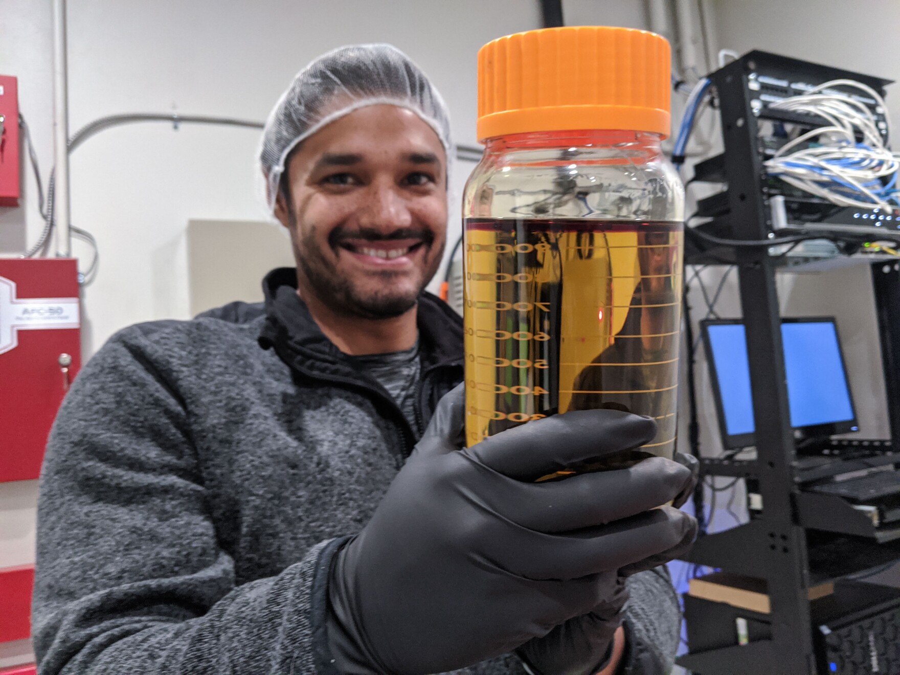 a smiling man wearing gloves and a hair net holds up a large glass jar filled with a yellow liquid