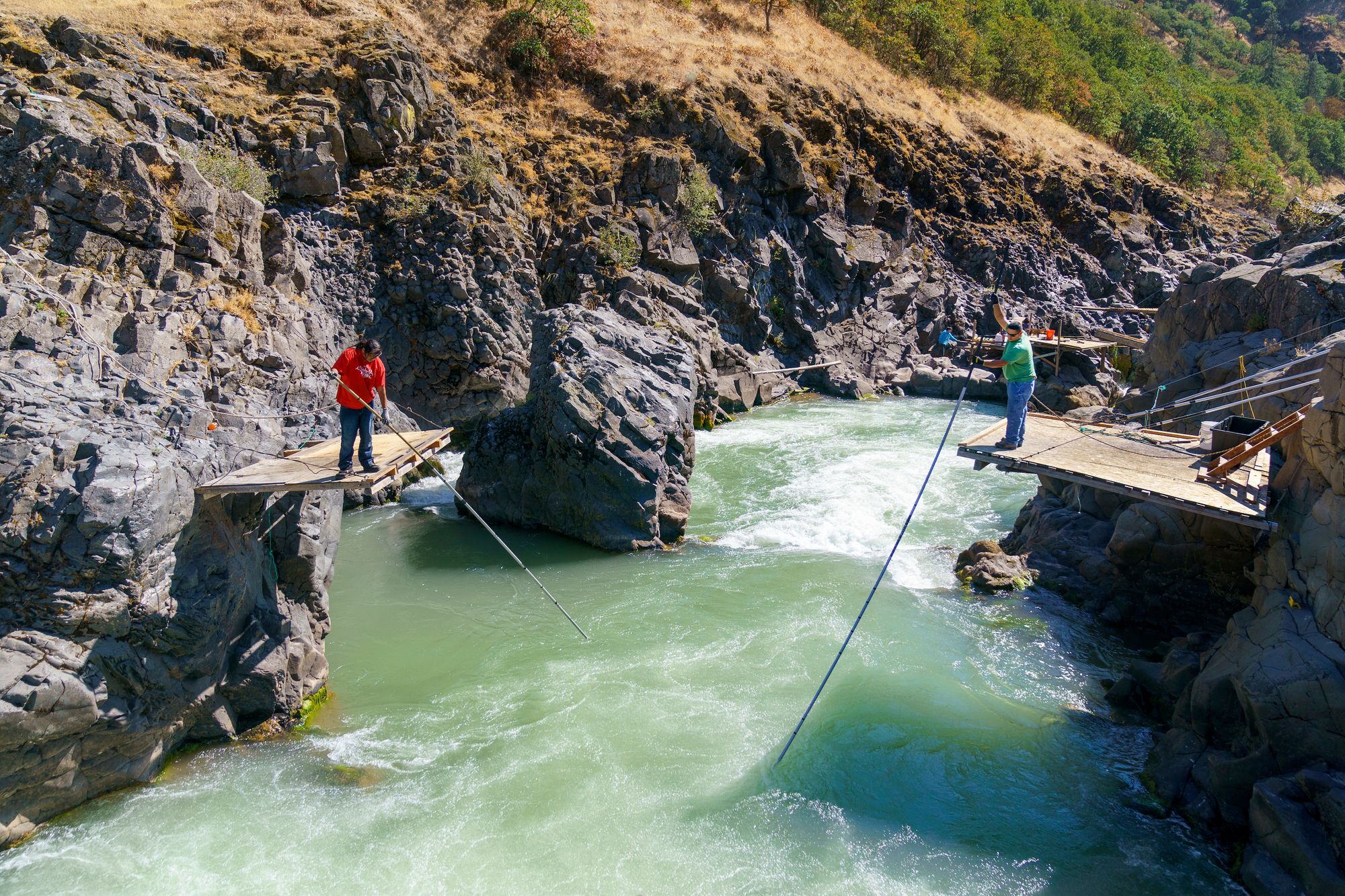 Two men stand on wooden platforms built into the rock above a river and extend long poles into the water.