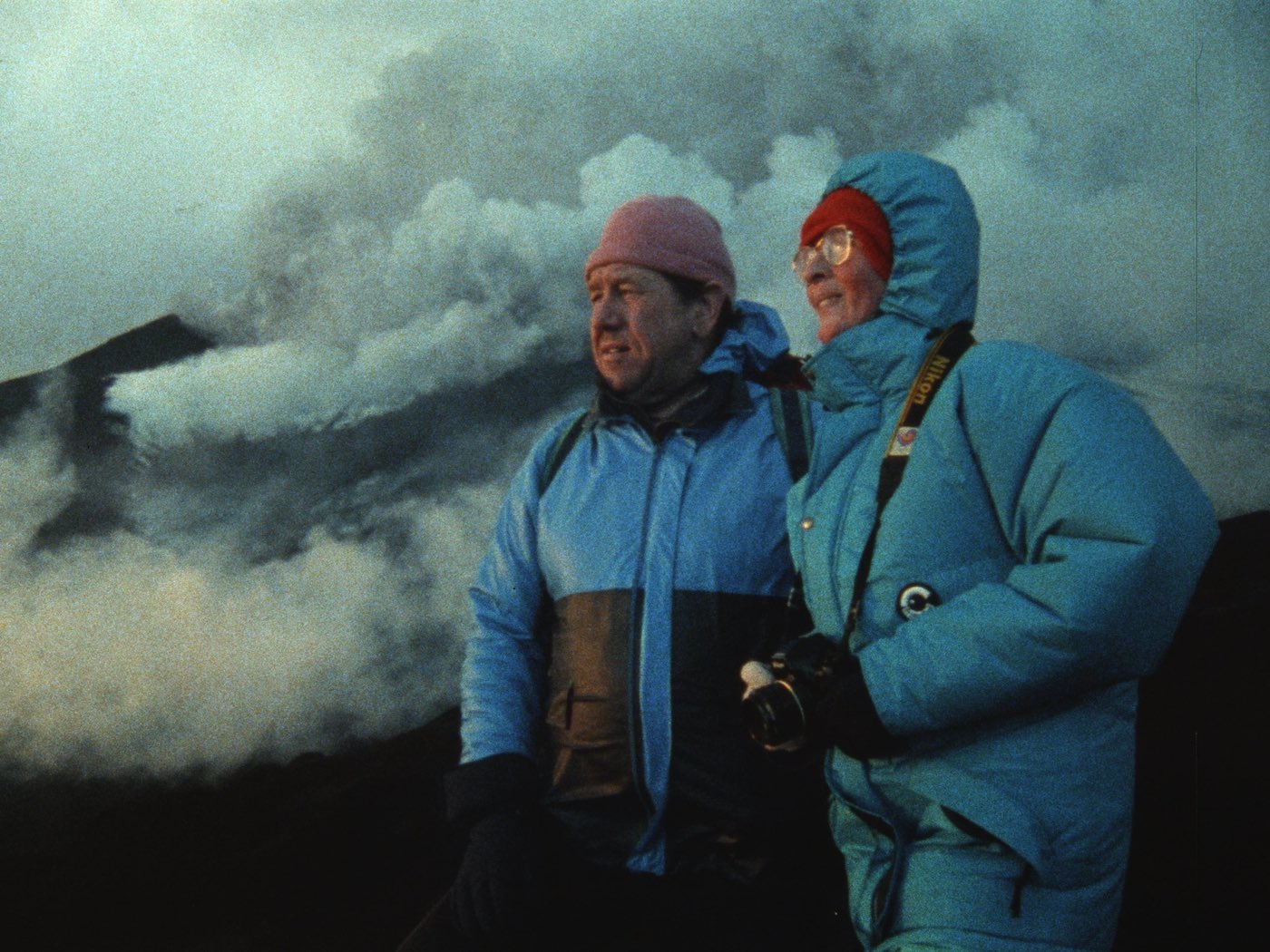 A man and a woman looking to the left of the frame, standing with a smoky hill, presumably a volcano in the background.