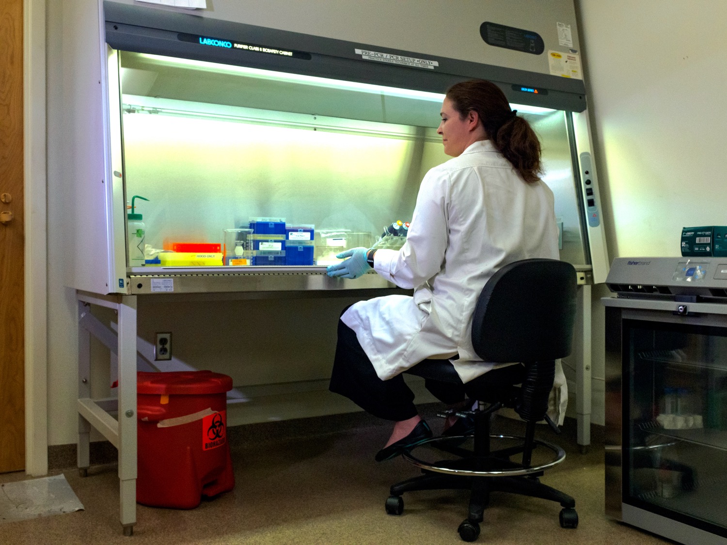 a white woman wearing a labcoat sitting down in a chair in a lab. in front of her is a large container with a glass front. scientific equipment is inside of it