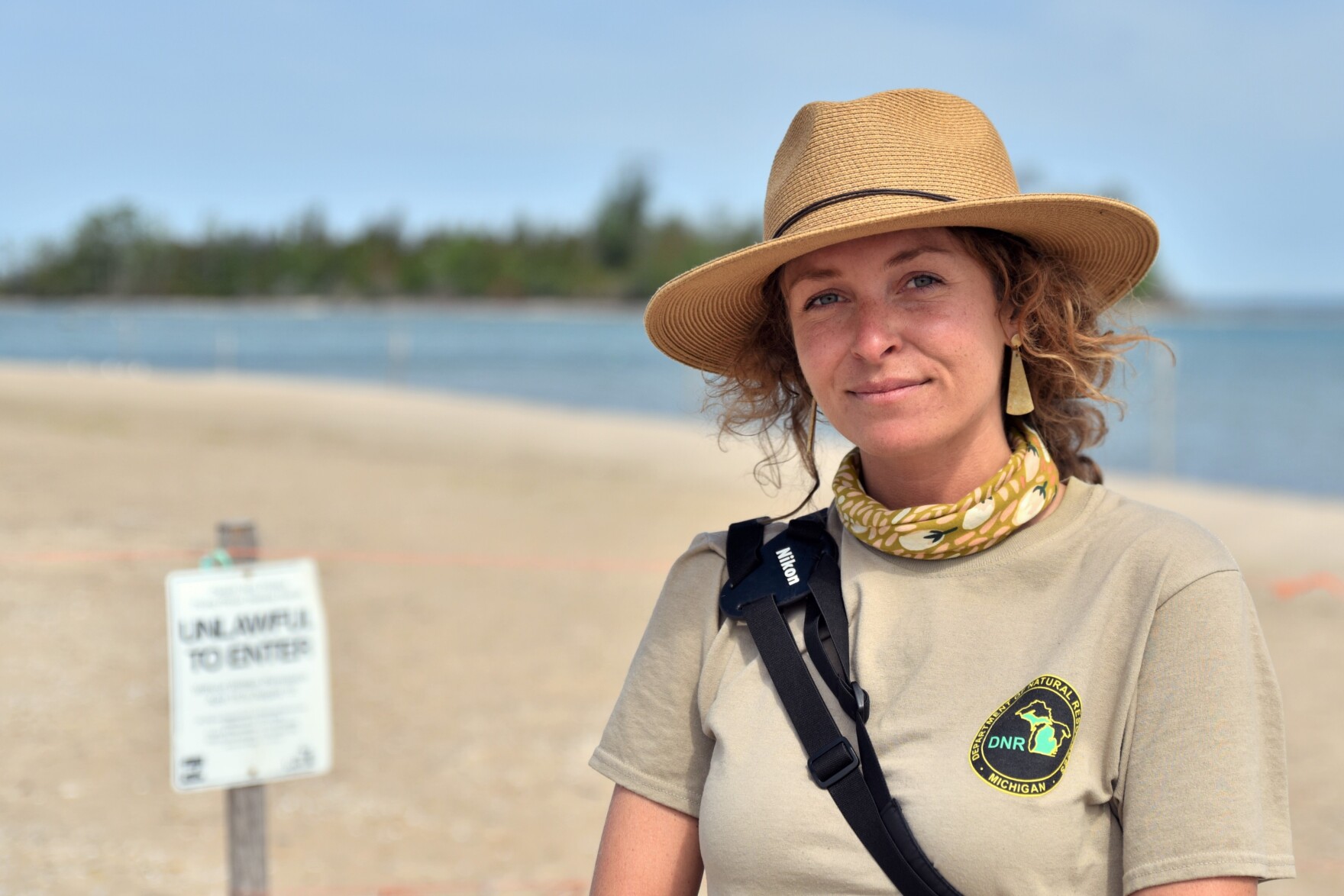 a young white woman standing on the beach with water behind her wearing a national park t-shirt and a wide brimmed hat smiles at the camera