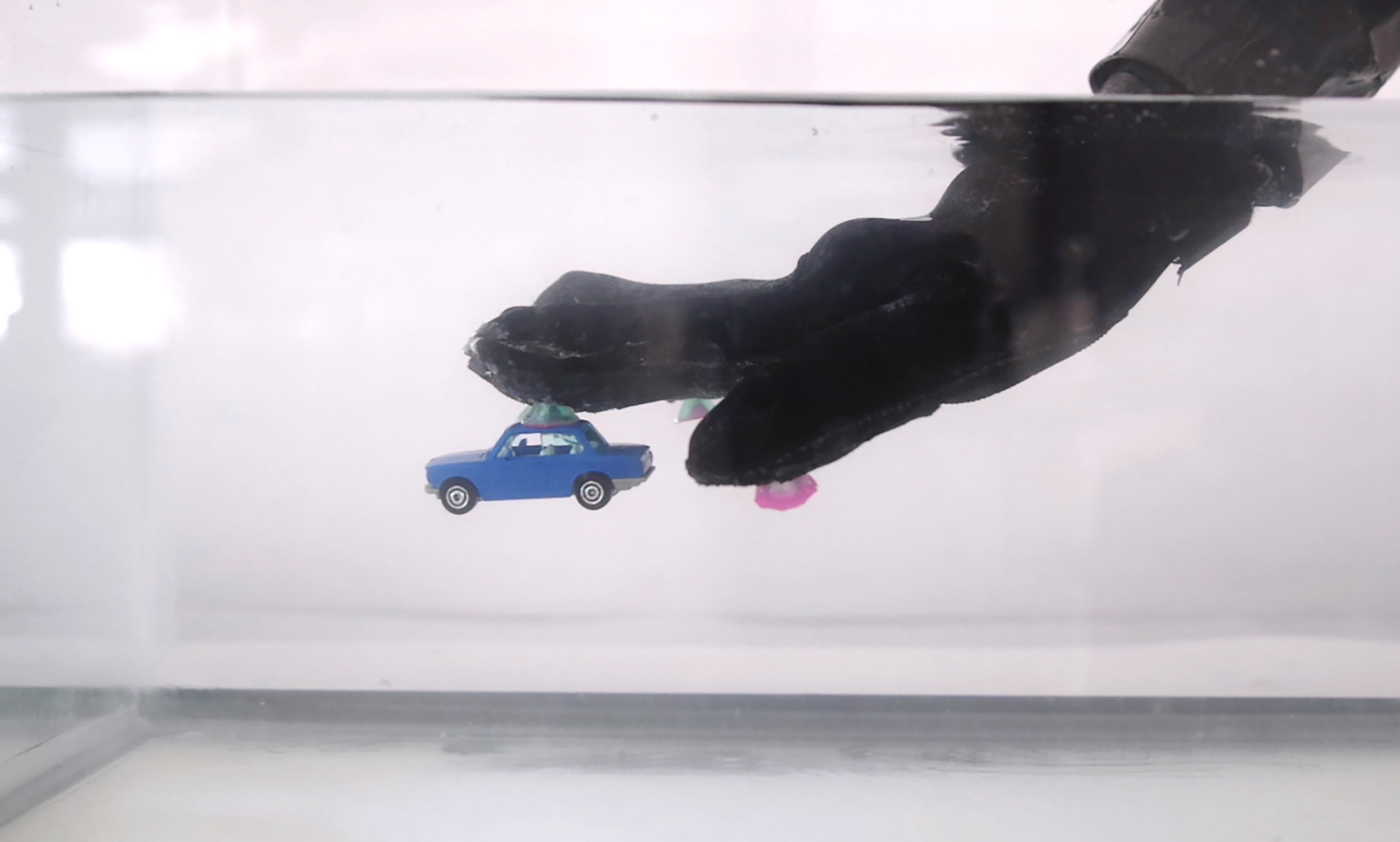 a side view of a two foot long tank of water. a large black glove reaches in and a small toy car somehow adheres to the bottom of the fingers
