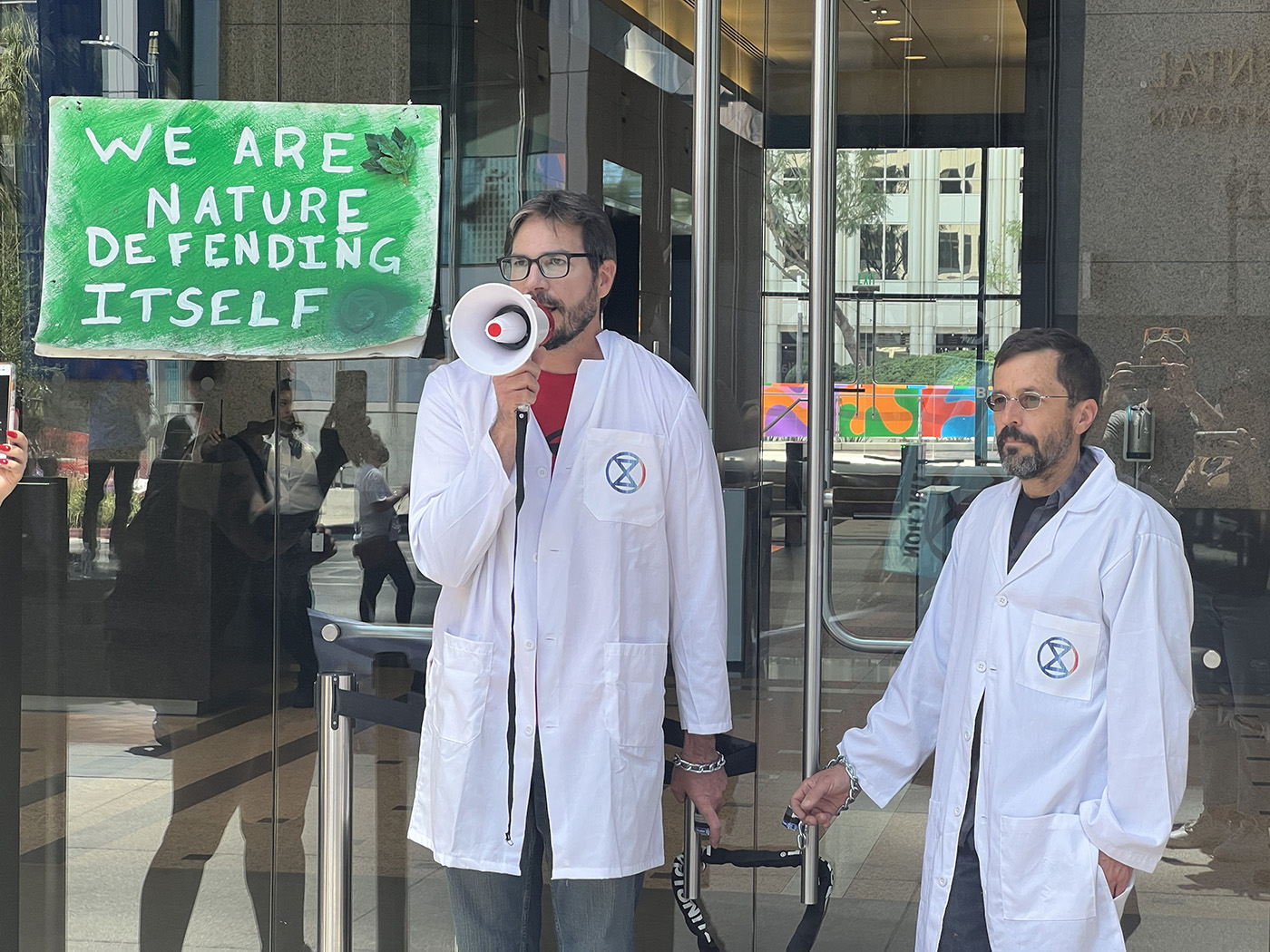 Two men chaining themselves to doors of a JPMorgan Chase & Co. Bank in Los Angeles. Behind them is a sign that reads: We are Nature Defending Itself 