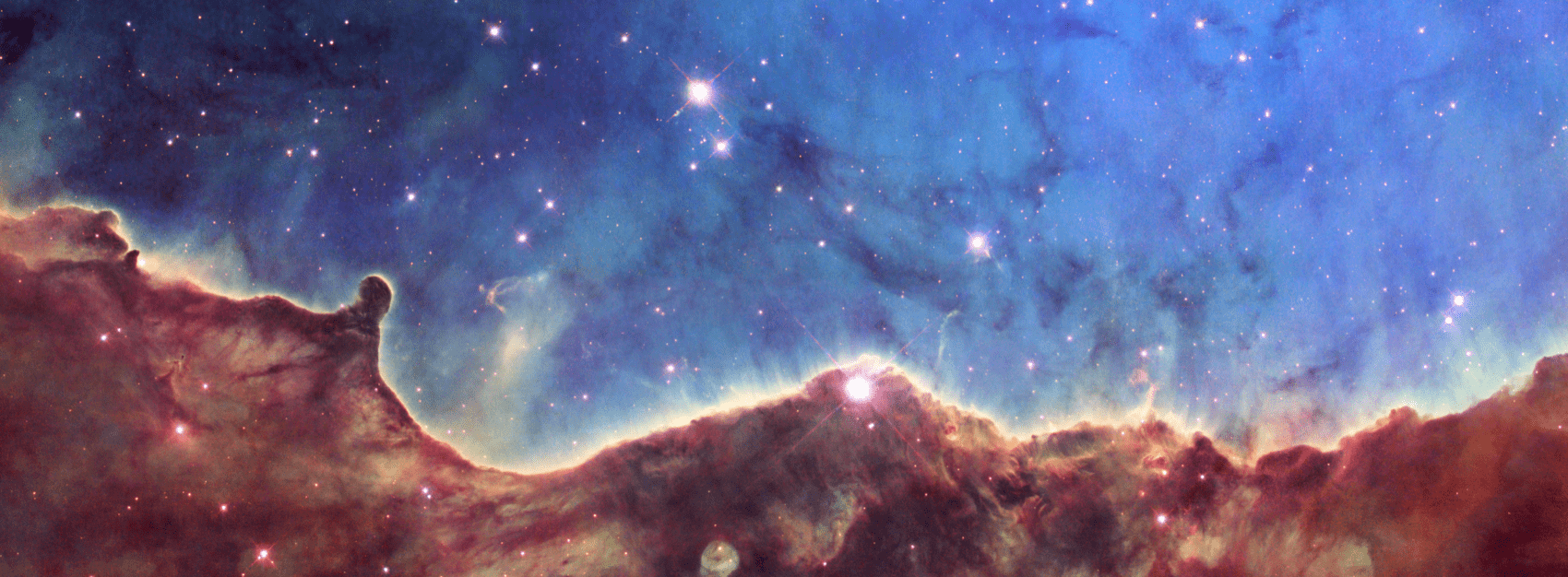 This Hubble image of the Carina Nebula shows the same region as a new Webb telescope image. Hills and valleys of gas and dust are displayed in intricate detail, cutting through a star-forming region. Wispy tendrils of gas, as well as dark trunks of dust, are set amid a backdrop of soft, glowing blue light. The glowing nebula has been carved out by intense ultraviolet radiation and stellar winds from several hot, young stars. The image also reveals dramatic dark towers of cool gas and dust that rise above the glowing wall of gas. The dense gas at the top resists the blistering ultraviolet radiation from the central stars, and creates a tower that points in the direction of the energy flow.