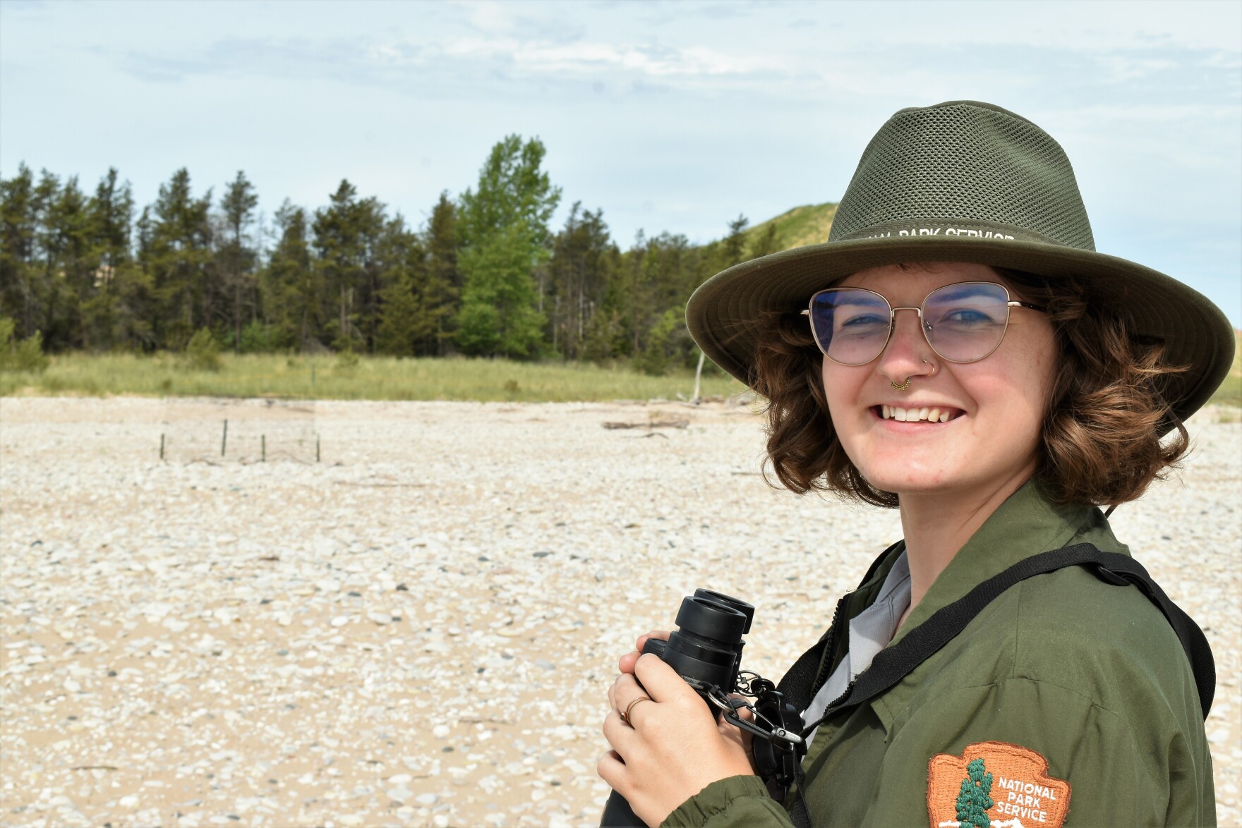 a younger white person wearing a national park service uniform smiles at the camera while holding bincolulars on the beach