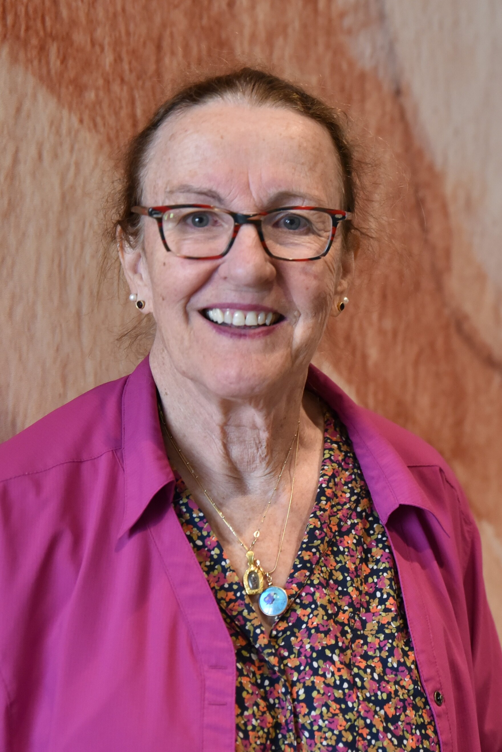 an older white woman wearing glasses, a pink shirt over a dress smiles at the camera