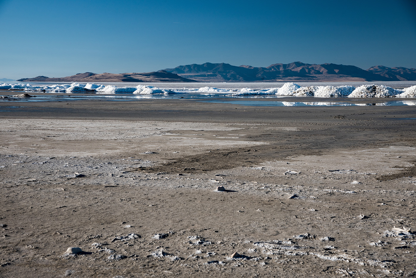 Dried, dusty, rocky ground on the lower two-thirds of the image. In the near distance, white mounds of salt pile high at the waterline of the lake. Utah's mountains rest in the far background.